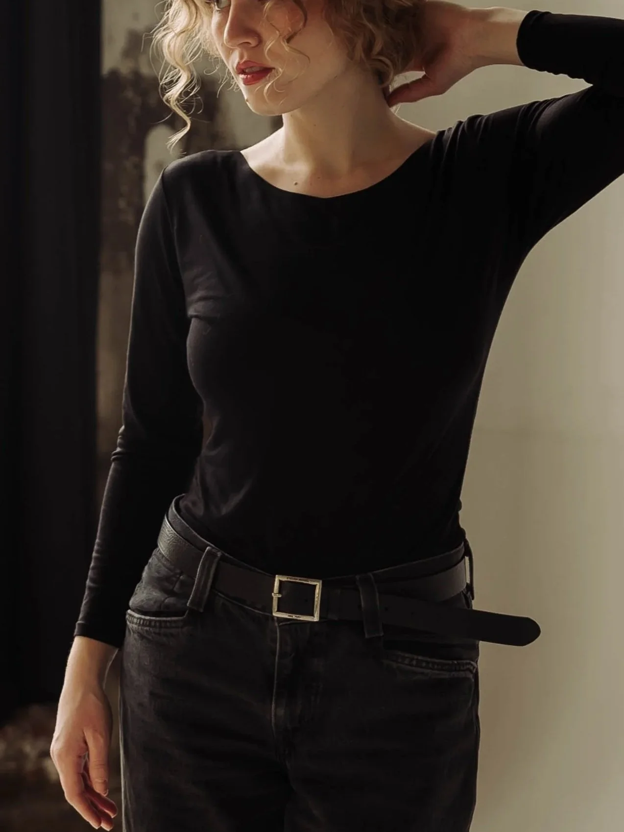A woman with curly blonde hair wearing a black long-sleeve shirt and black jeans with a belt. She has her left hand behind her neck and is holding a black object with her right hand, standing against a blurred background.
