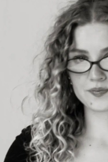 A woman with curly hair and glasses, wearing a dark top, looking slightly to the side.