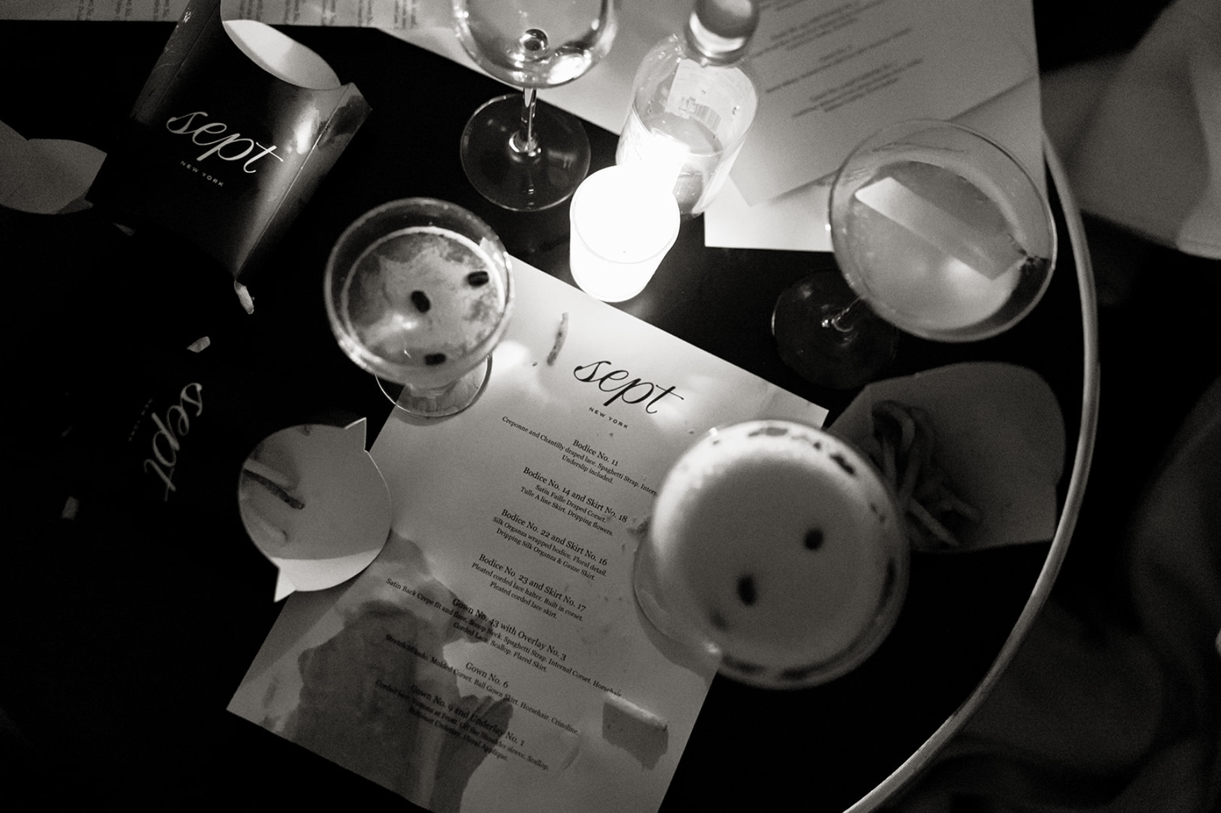 A black-and-white photo showing a table with several glasses and a lit candle, with a 'sepot' New York menu or program placed underneath.