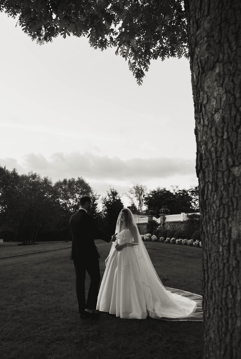 A black and white photo of a bride and groom outdoors under a tree during a wedding ceremony, with the couple holding hands and facing each other on a grassy area.