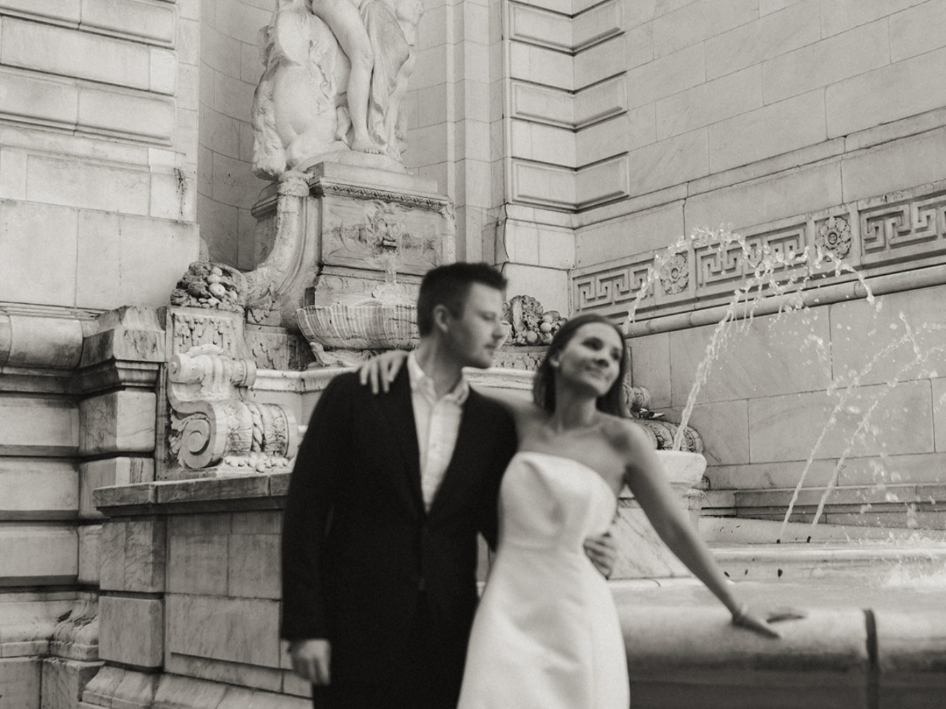 A black-and-white photo of a man and woman in elegant attire standing next to a fountain with ornate sculptures, water flowing from the fountain, in a marble architectural setting.