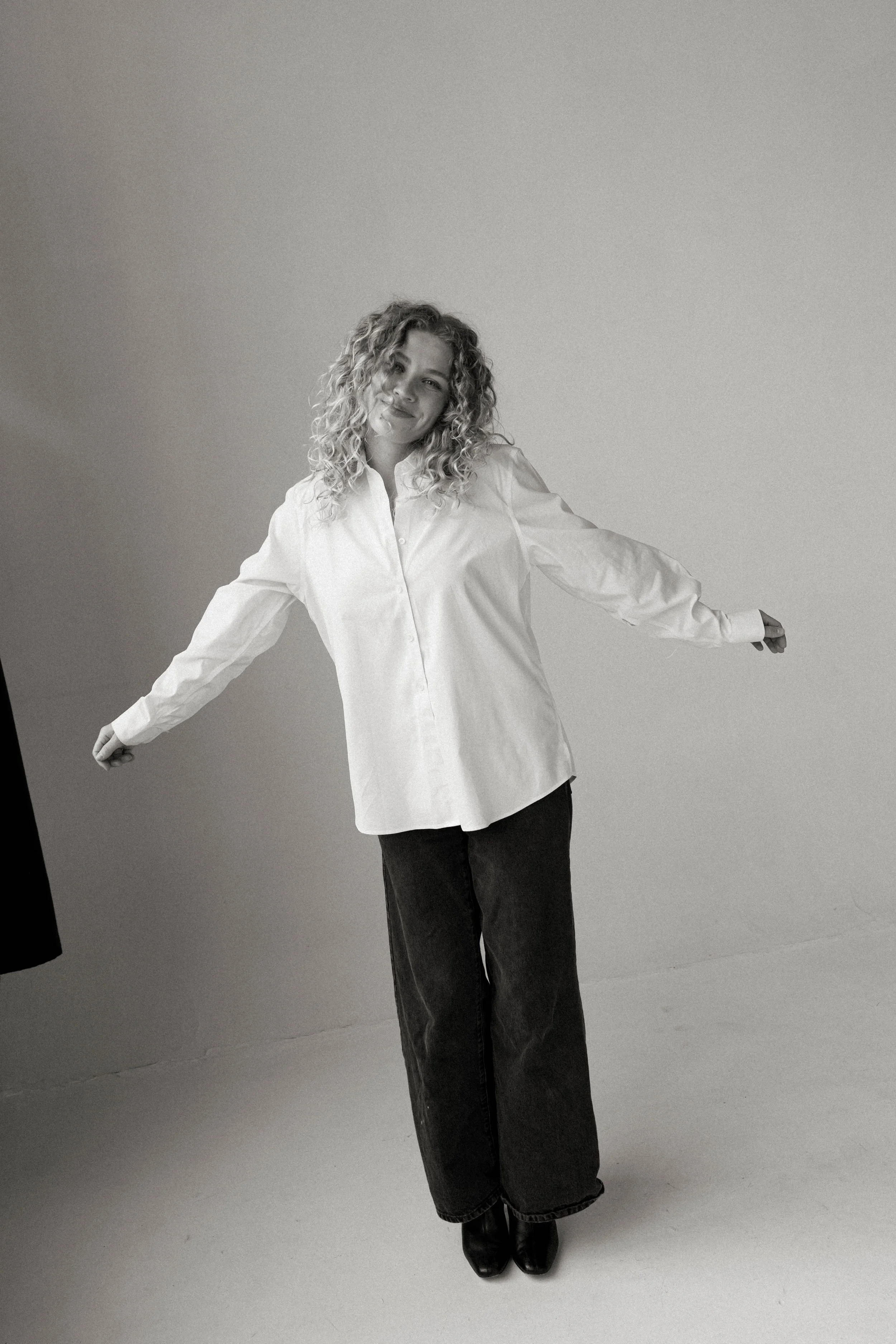 A black and white photo of a smiling woman with curly hair, wearing a loose white shirt and dark pants, standing with her arms outstretched against a plain background.