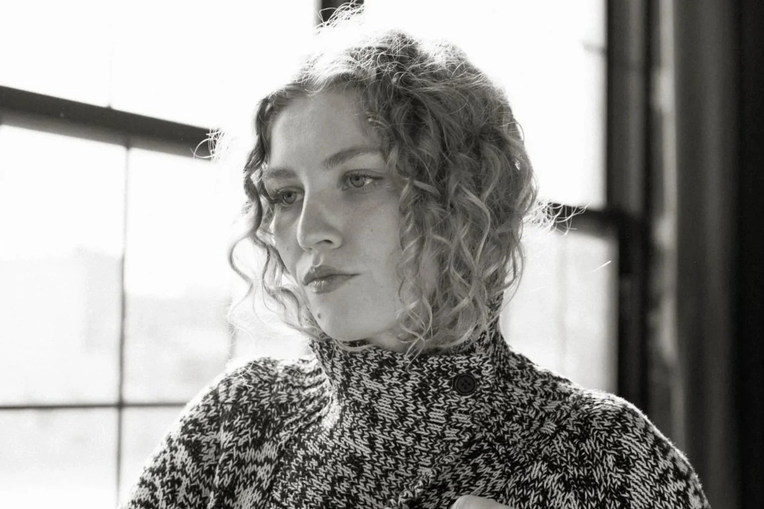 Black and white portrait of a woman with curly hair and a patterned sweater, looking contemplative, near a window with light streaming in.