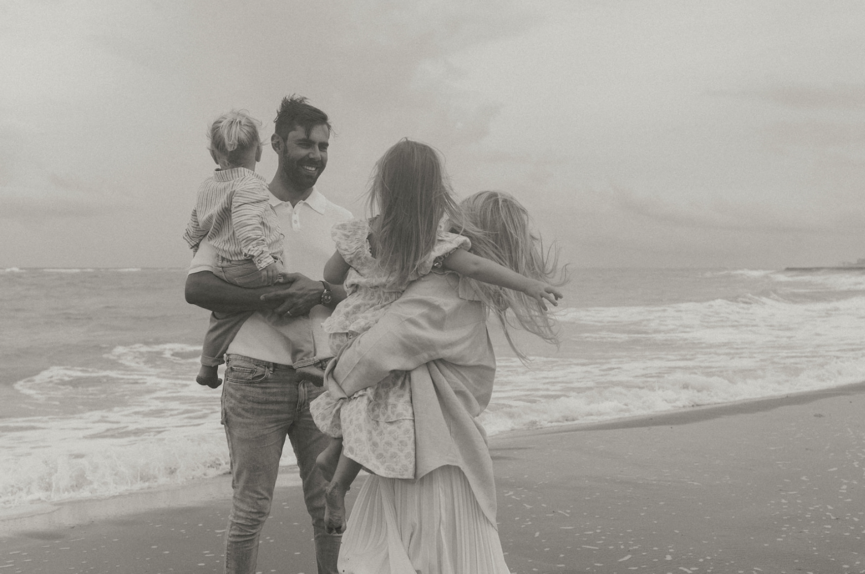 A family enjoying time together on the beach, with two adults carrying two children, all smiling and interacting near the shoreline.