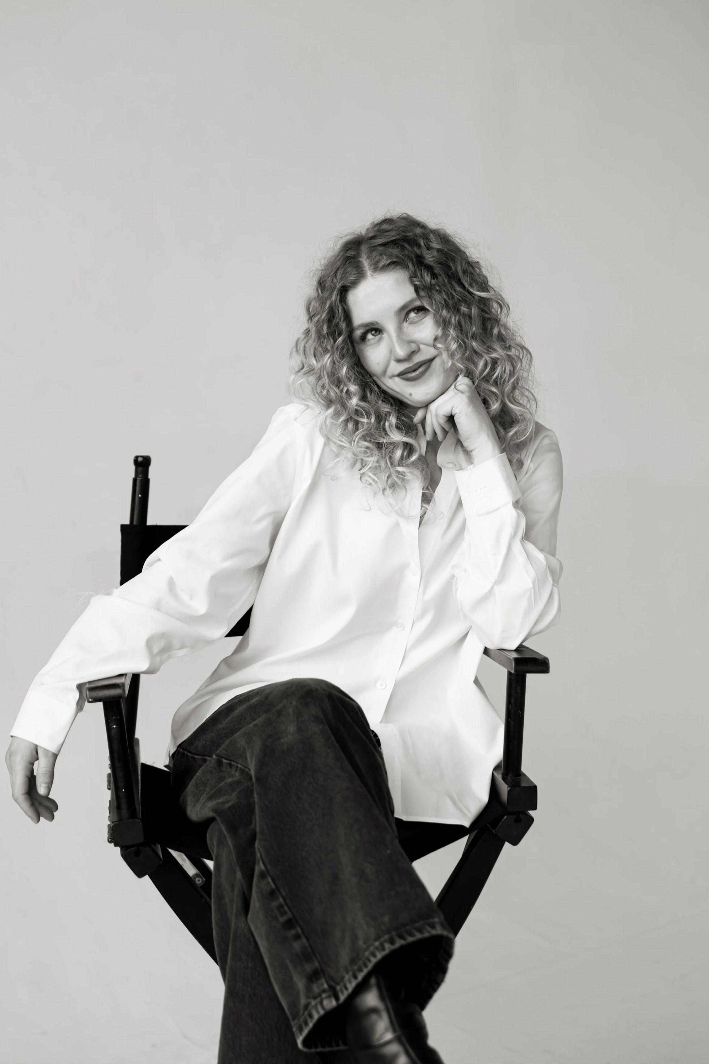 Black and white photo of a woman with curly hair sitting in a director's chair, wearing a white shirt and dark pants, smiling thoughtfully.