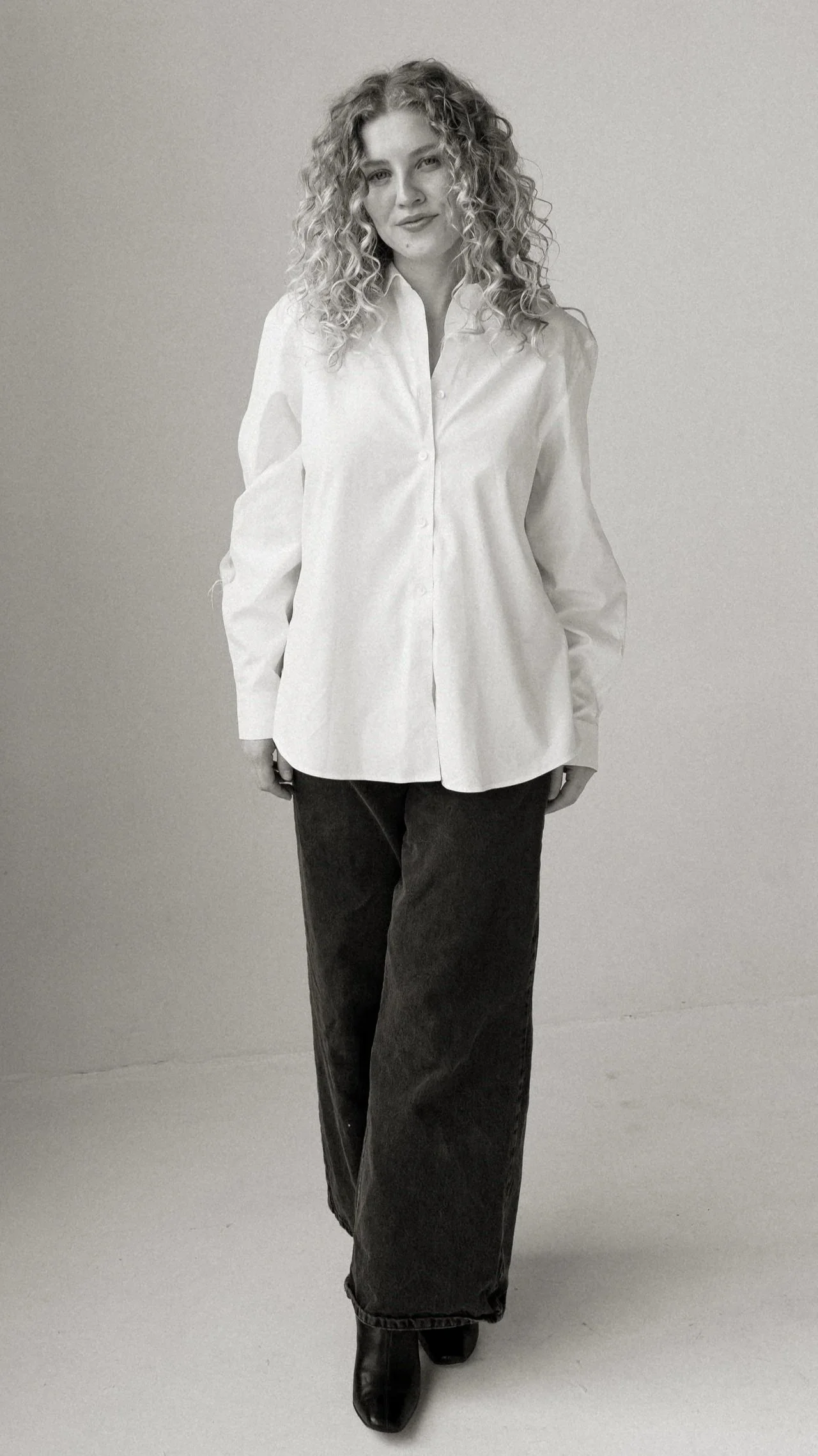 A woman with curly hair wearing a white button-up shirt, dark pants, and boots, standing against a plain background.