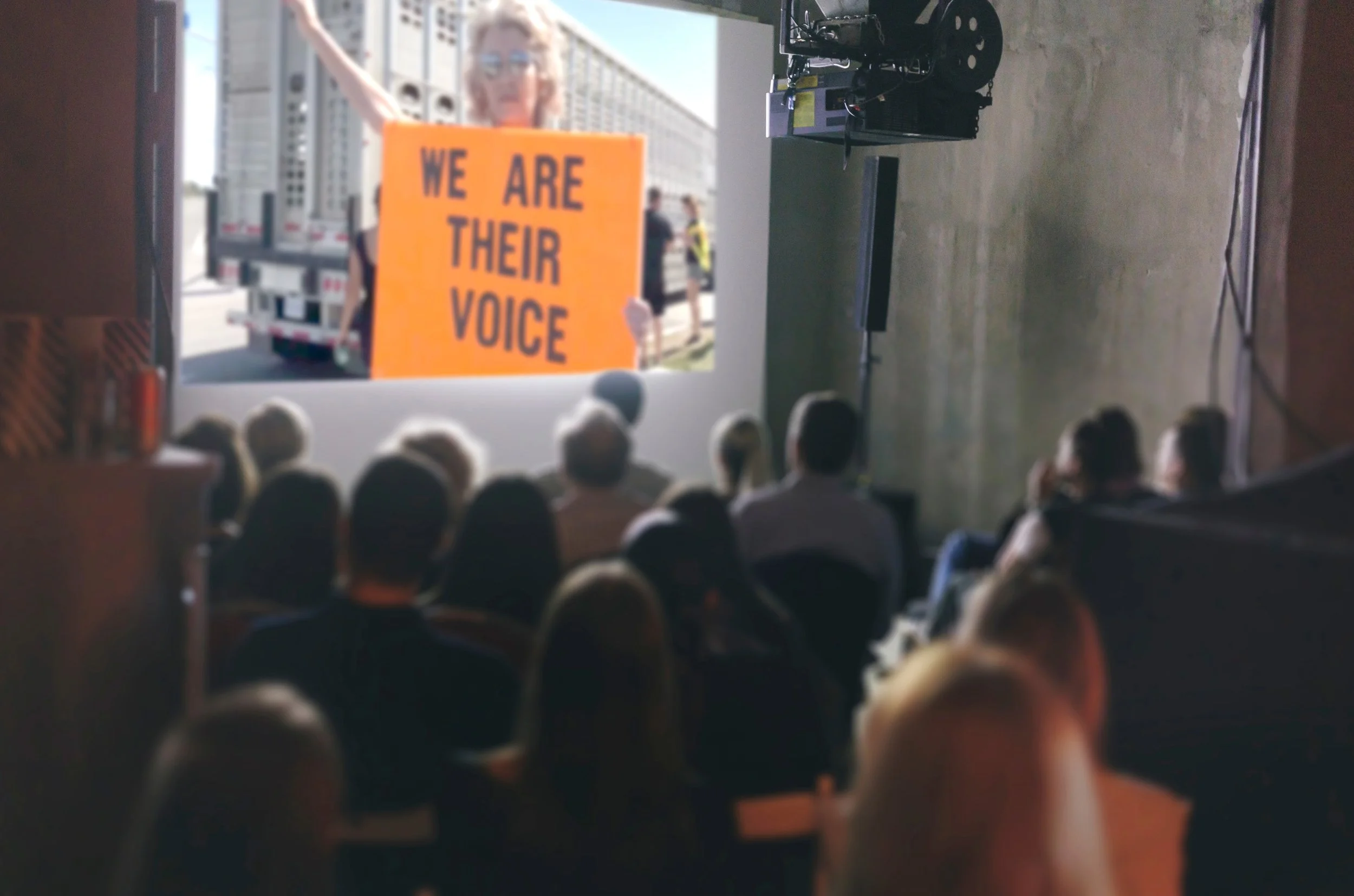 A group of people watching a video screen inside a room with concrete walls. The video shows a woman holding a sign that reads 'WE ARE THEIR VOICE,' and she appears to be at an outdoor protest or demonstration.