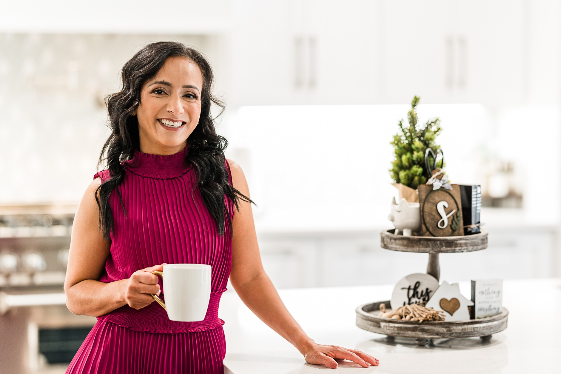 Smiling woman in a pink dress holding a white mug in a bright kitchen with holiday decorations on a tiered tray to her right.