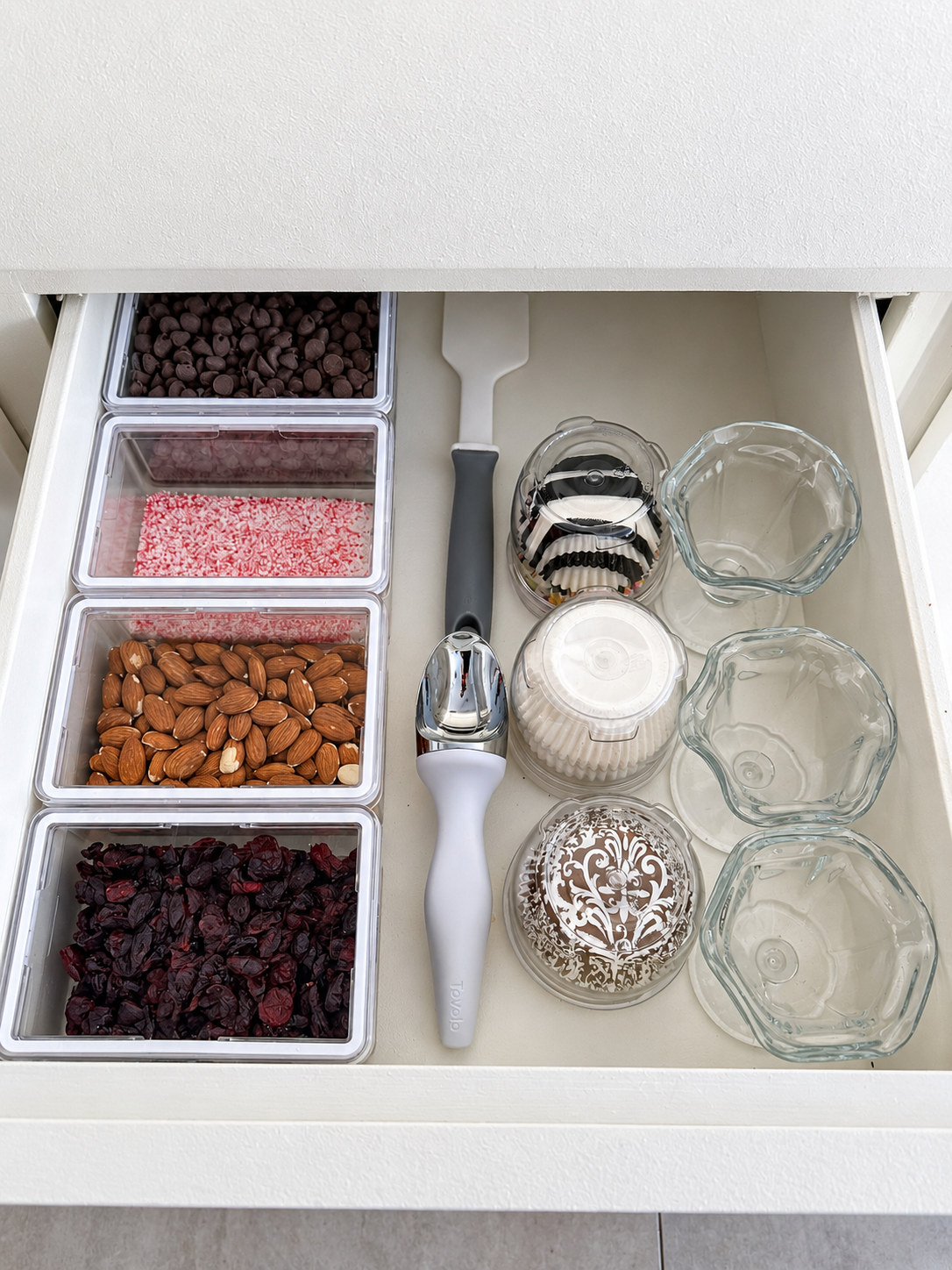 four clear containers with lids showing nuts, cranberries, peppermint and chocolate chips, an ice cream scooper, paper cupcake cups, and clear parfait cups.