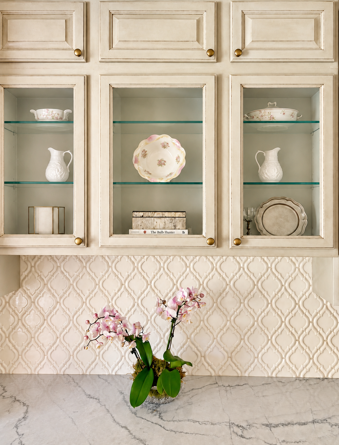 cream cabinetry with china display above a marble countertop with a pink orchid.