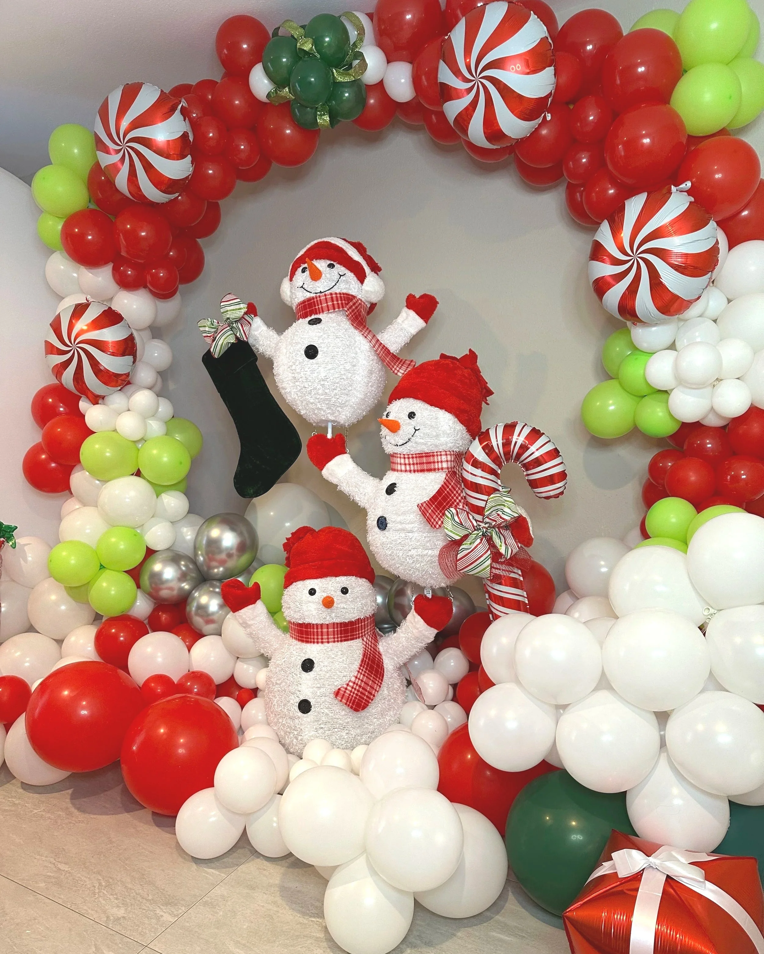 Christmas display with three snowmen framed by a red, white, and green balloon arch with peppermint decorations.