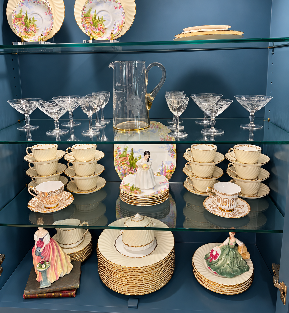 Crystal glasses and pitcher, cream china plates and tea set with gold trim. Some plates have flowers. and 3 small female figurines styled to look like a store display.