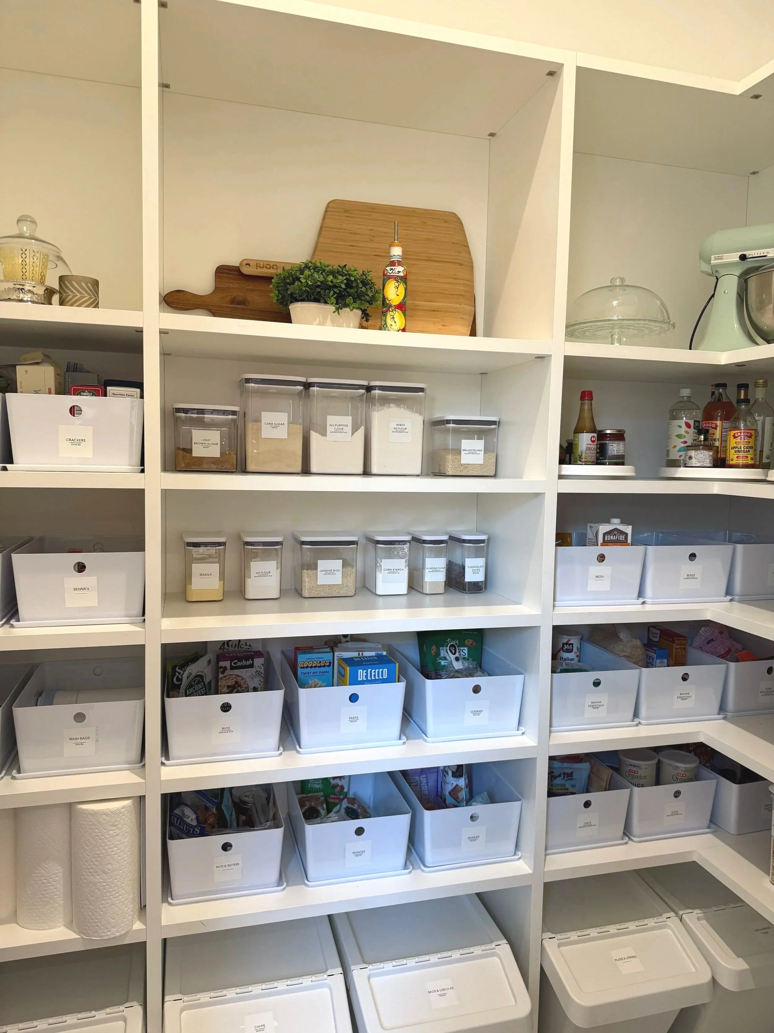 White pantry shelves with labeled food containers, grouped baking supplies, cutting boards, and decorative accents.