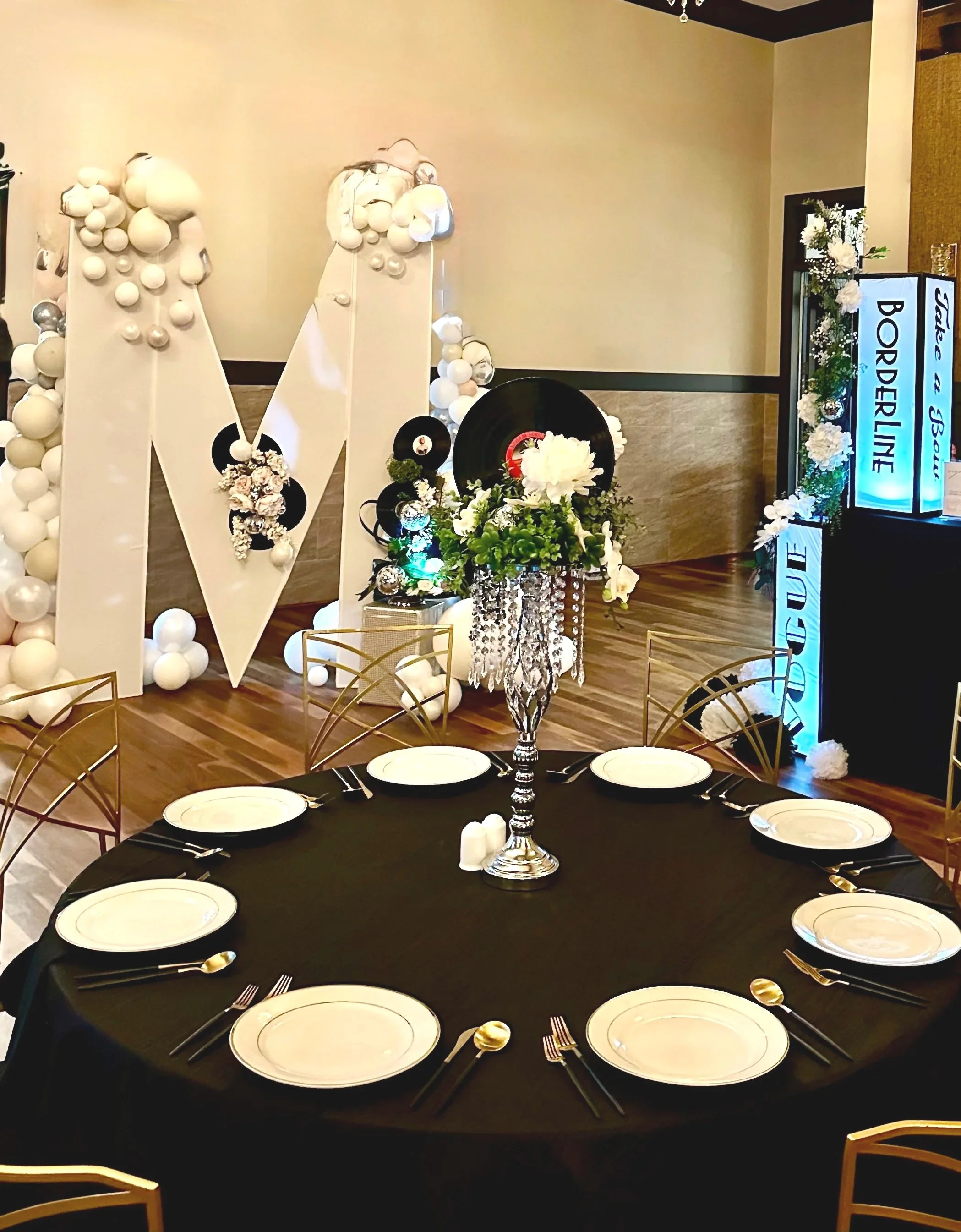 Black table setting with white plates, gold chairs, and a tall floral centerpiece with hanging crystal strands, beside a balloon-decorated white letter M.