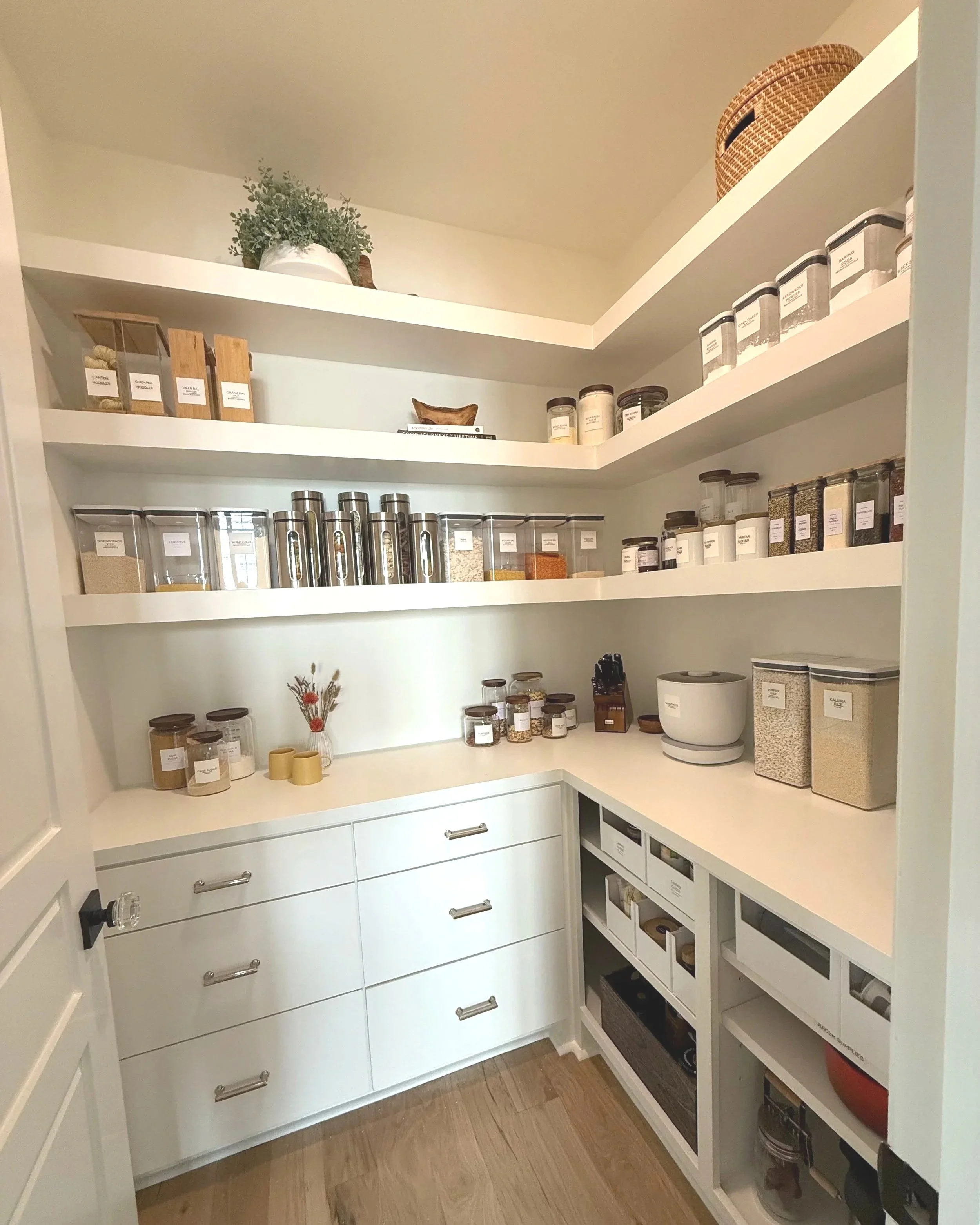 Walk-in pantry with white shelves and drawers, labeled food containers, and grouped kitchen supplies.