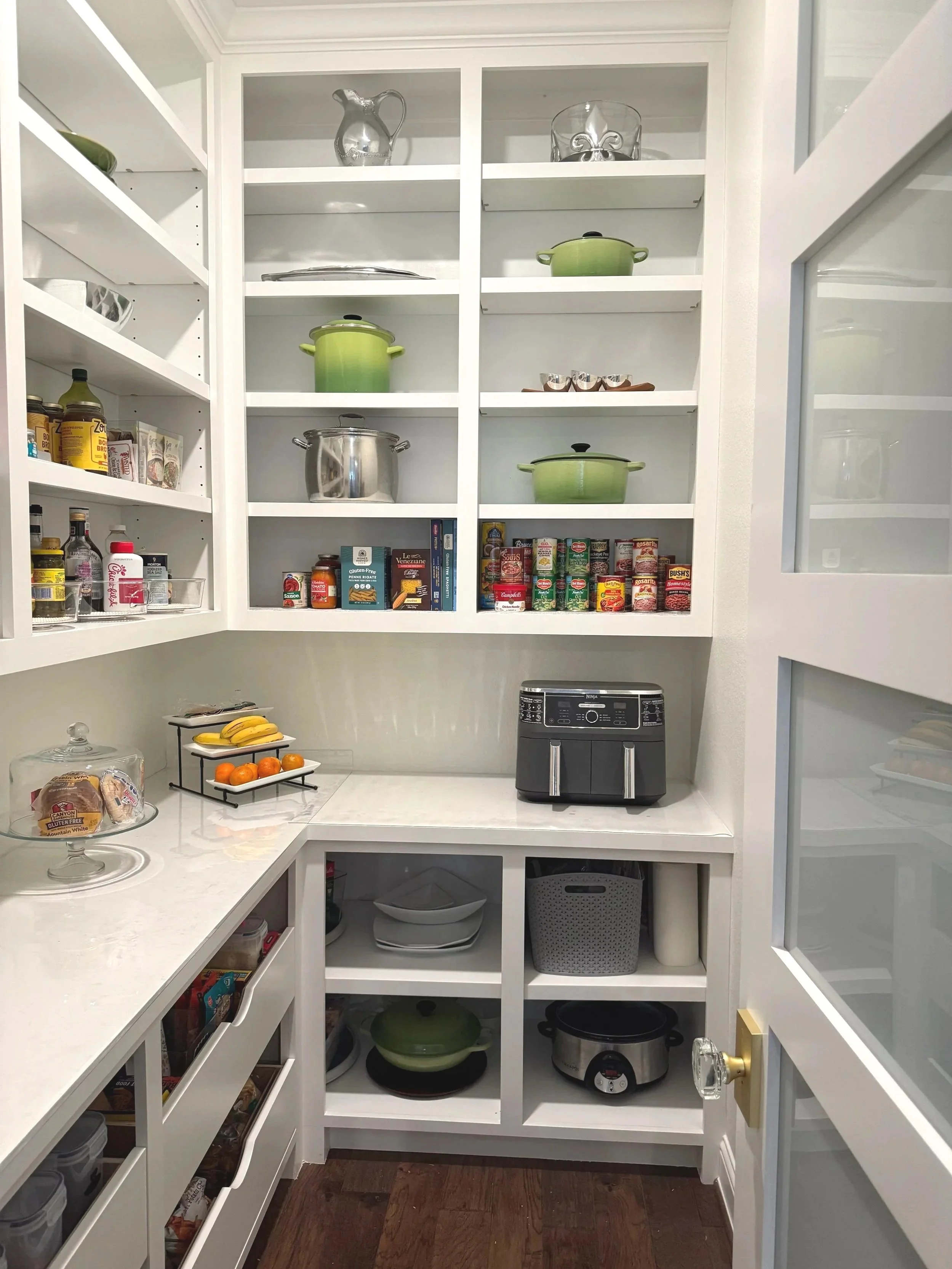 Inside a pantry with white shelves filled with cookware, canned goods, and other kitchen items. There are green pots on the shelves, a rice cooker, bread, bananas, and a bowl of oranges on the countertop.