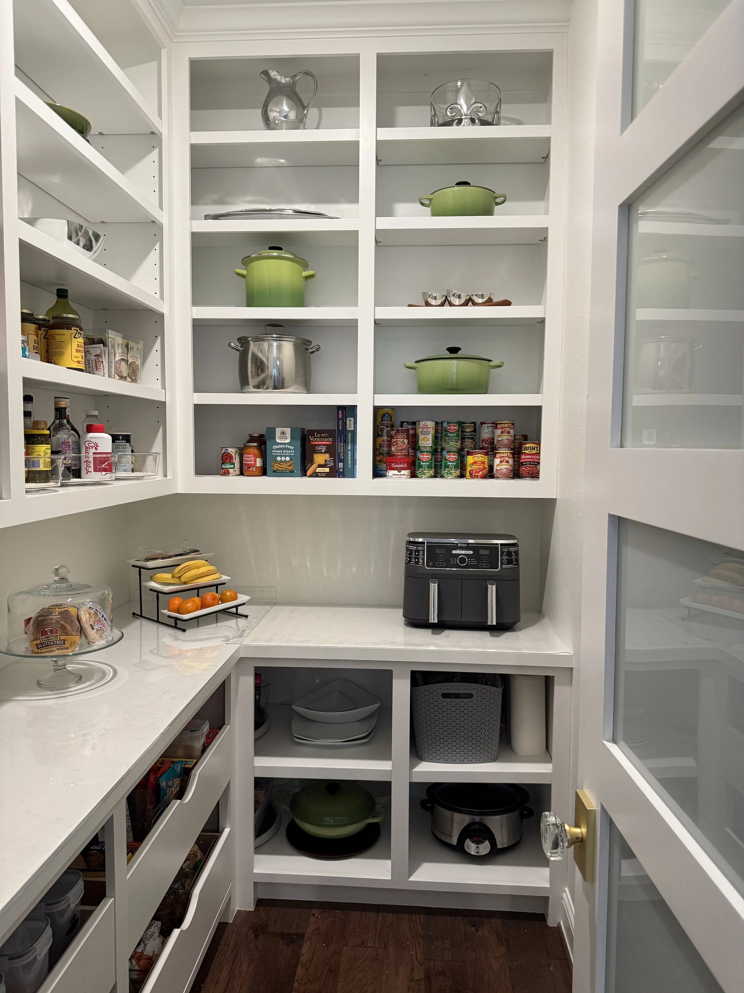 A pantry with white shelves filled with kitchenware, canned goods, and food items. There is a small counter with bananas, oranges, and bread. Kitchen appliances like a slow cooker and an air fryer are visible.
