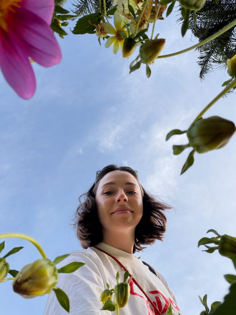 A woman taking a selfie outdoors, looking down at the camera, surrounded by blooming flowers and a blue sky with some clouds.