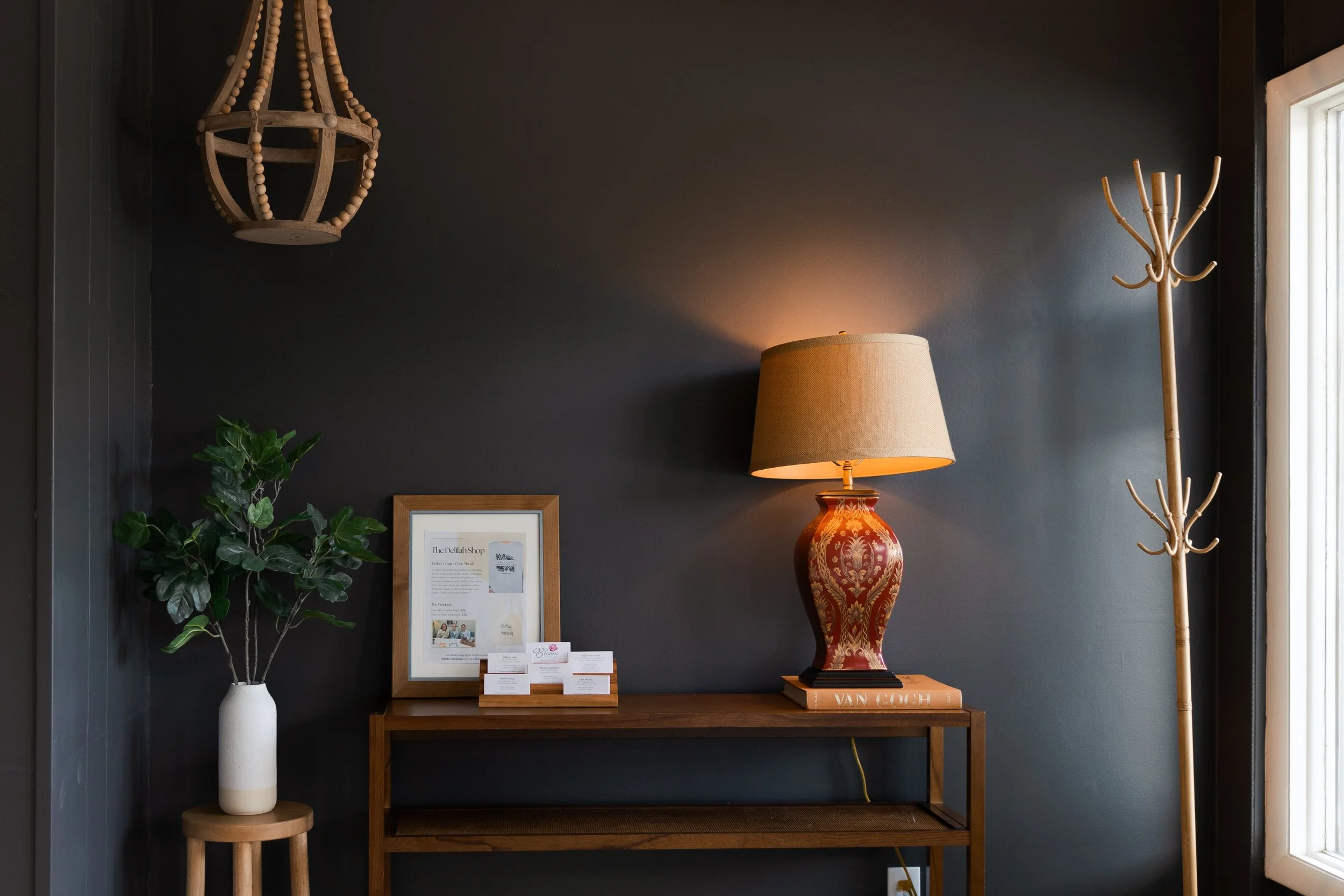 Interior of a room with a dark wall, wooden console table, decorative lamp, potted plant, business cards, framed paper, and a wooden coat rack near a window.