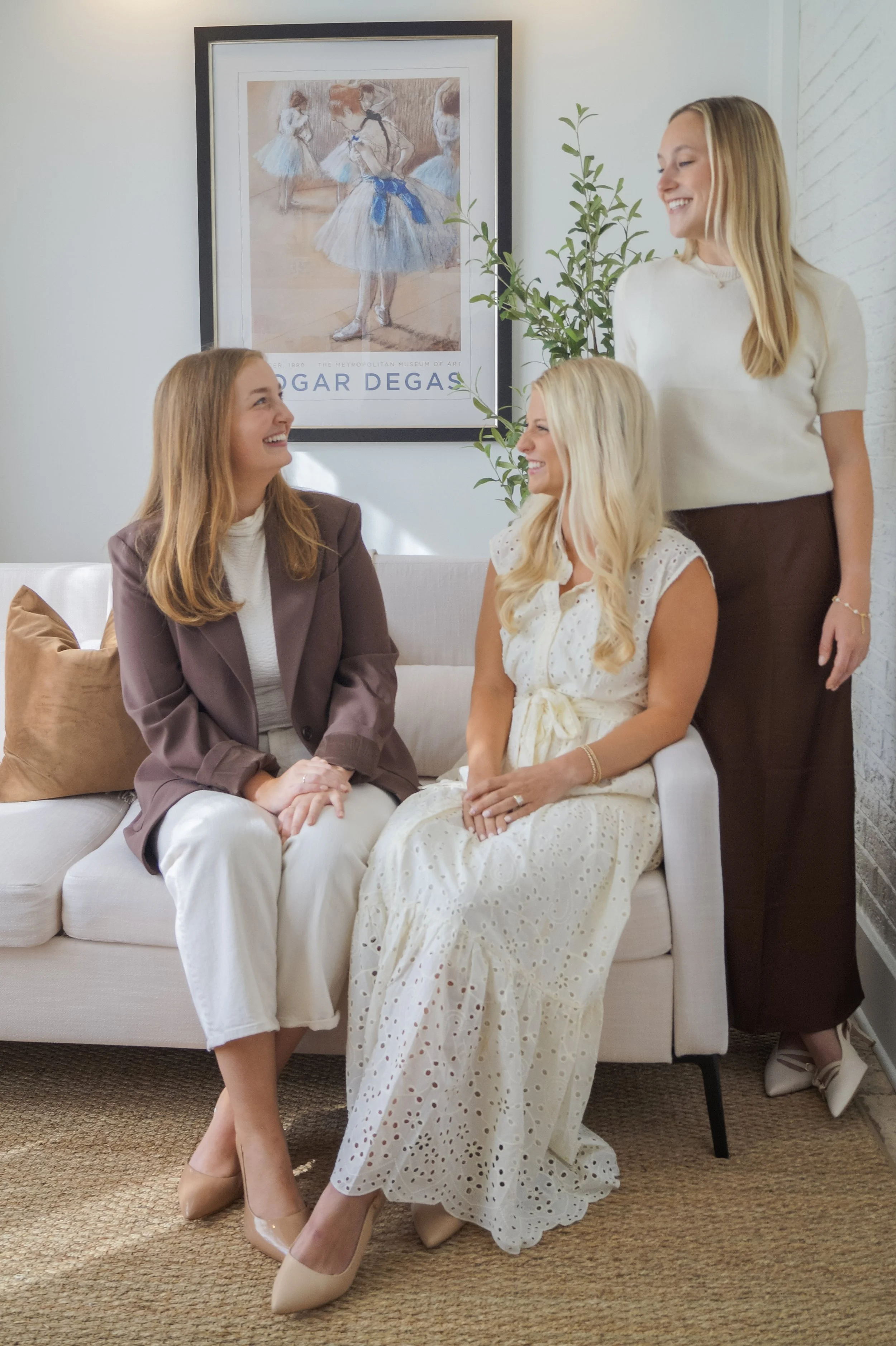 Four women with blonde hair and light skin smiling and talking in a living room. One woman is standing and the other three are sitting on a white sofa. The woman standing wears a cream-colored top and dark skirt. The woman sitting on the left wears a brown blazer and cream trousers. The woman in the middle wears a white dress with eyelet pattern. The woman sitting on the right has ginger hair and wears a white top. There is a framed ballet drawing on the wall behind them and a potted plant beside it.