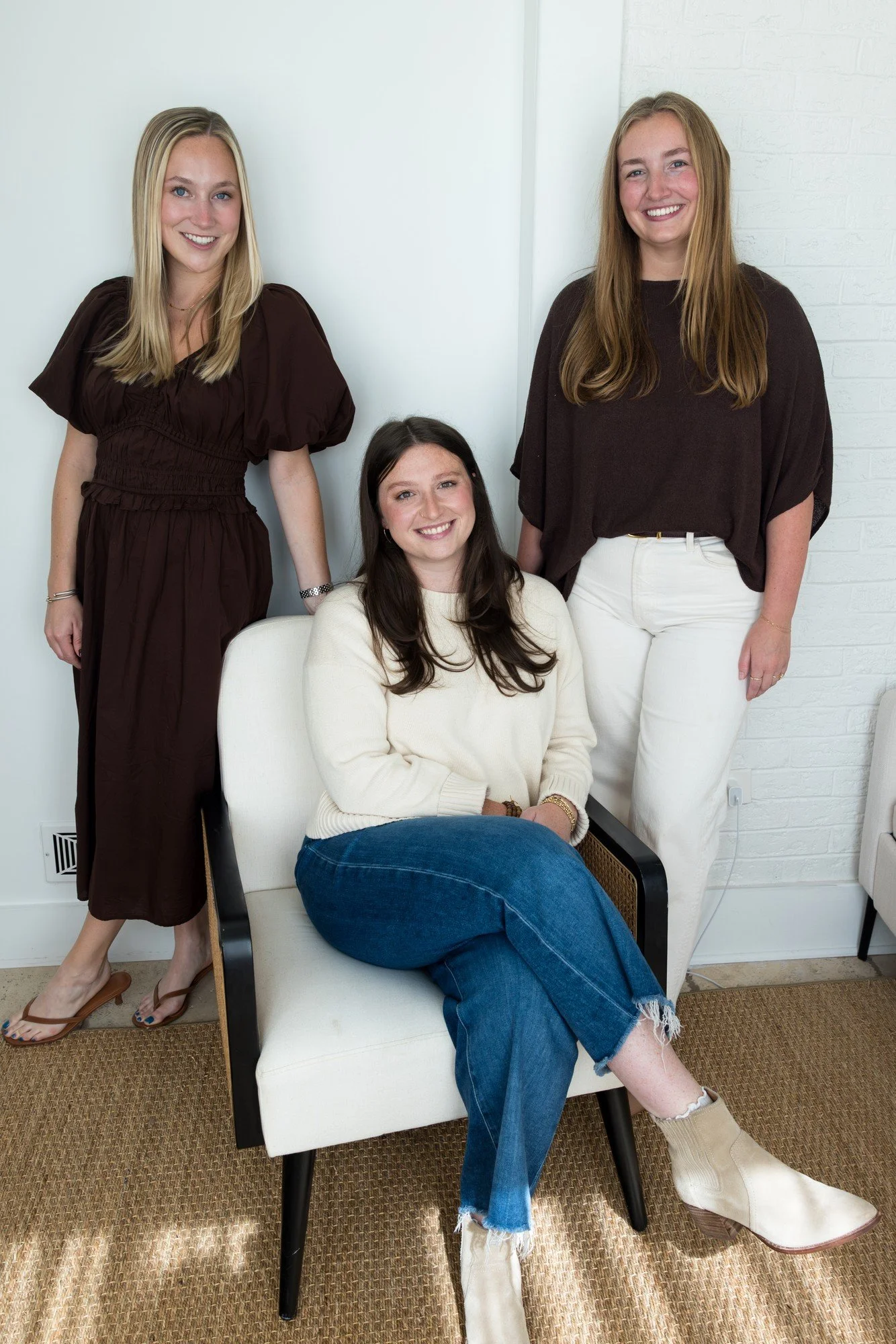 Five women sitting on a beige bench in a cozy, well-lit room, smiling and engaging with each other. They are dressed in stylish, semi-formal attire, with some wearing heels and others in boots. The room has a white brick wall on the left, a framed floral artwork above them, and a tall green potted plant on the right.
