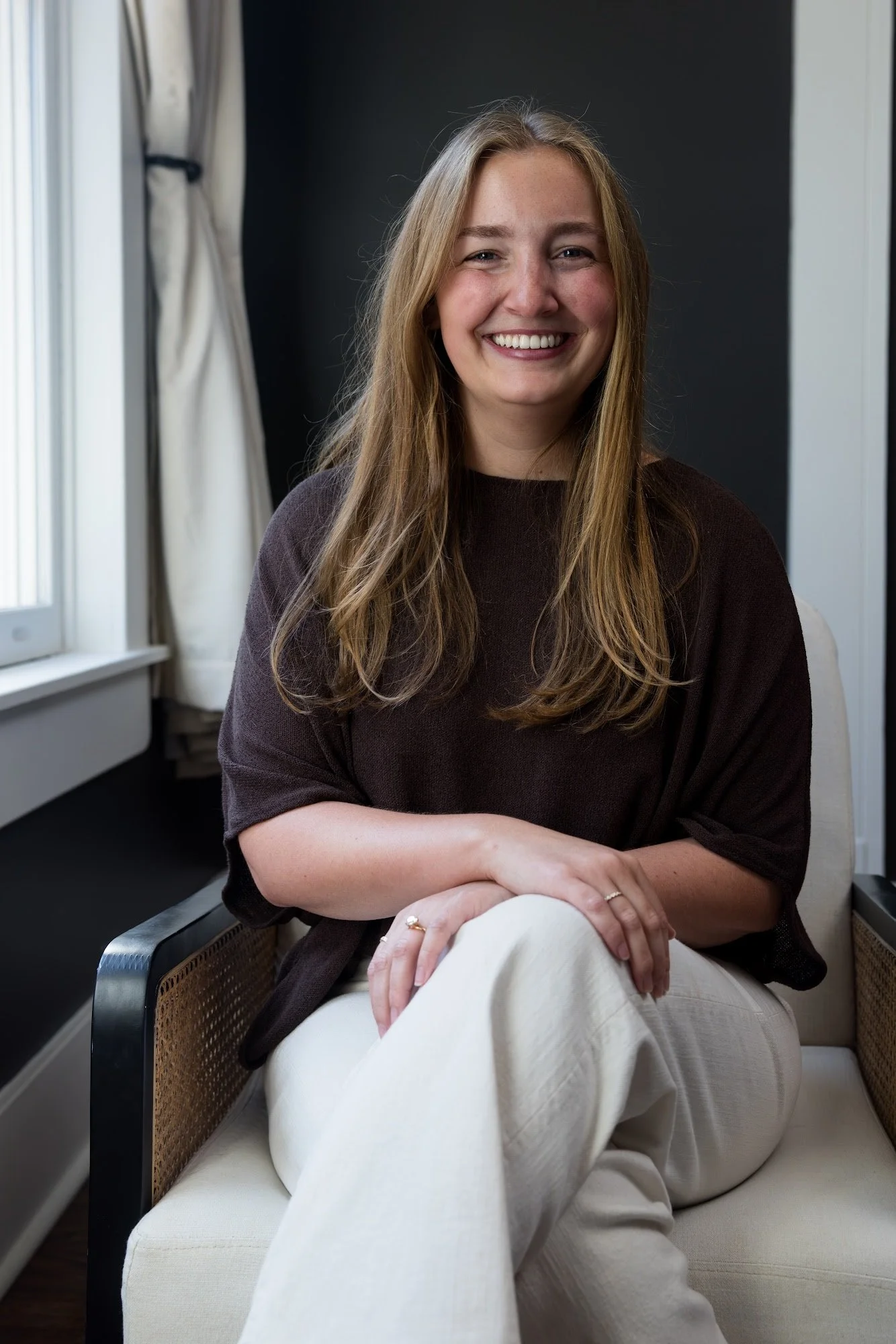 A young woman with long, blonde hair sitting on a light-colored sofa in a room with natural light. She is smiling, wearing a dark brown top and beige pants, with one arm resting on her knee and the other crossed over her lap. There is a window with curtains behind her.