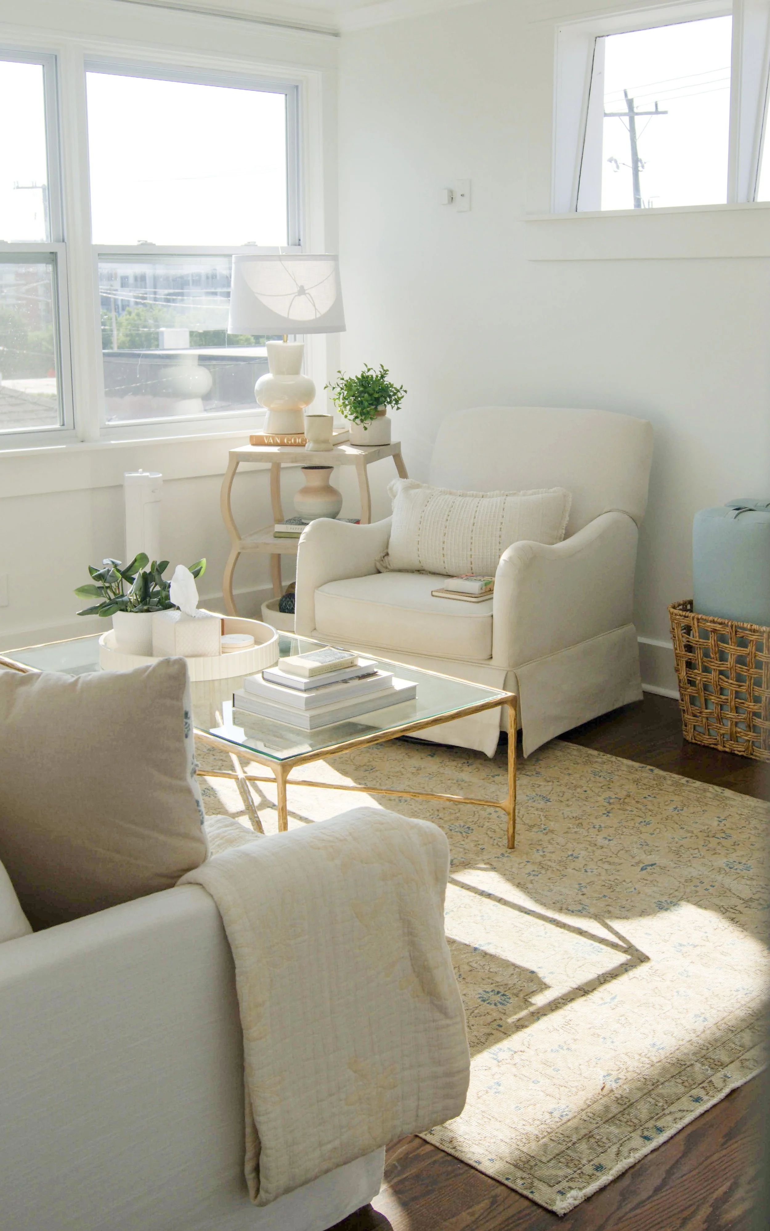 Bright living room with white sofa, armchair, gold coffee table with books, a side table with a lamp, plants, and a large window letting in natural light.