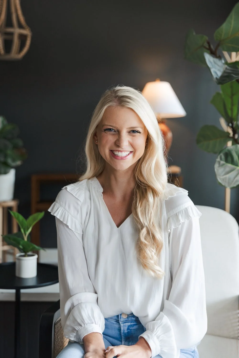 A smiling blonde woman wearing a white blouse and jeans sitting in a living room with indoor plants, a lamp, and dark-colored walls.