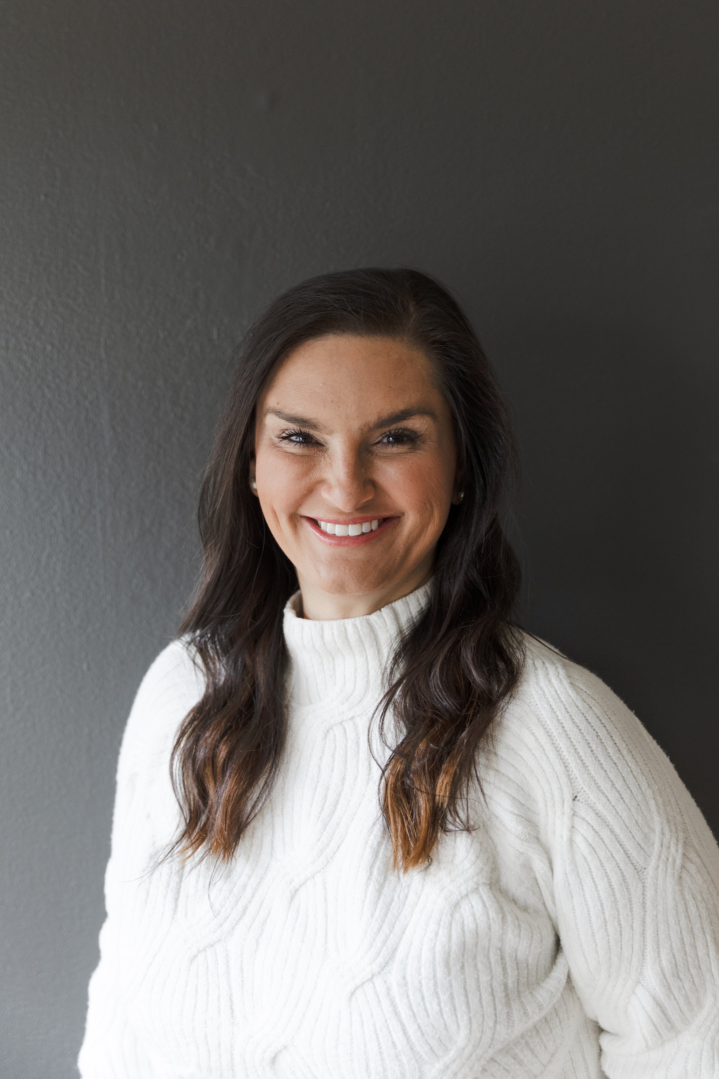 A smiling woman with dark brown hair wearing a white knitted sweater, standing in front of a plain dark background.