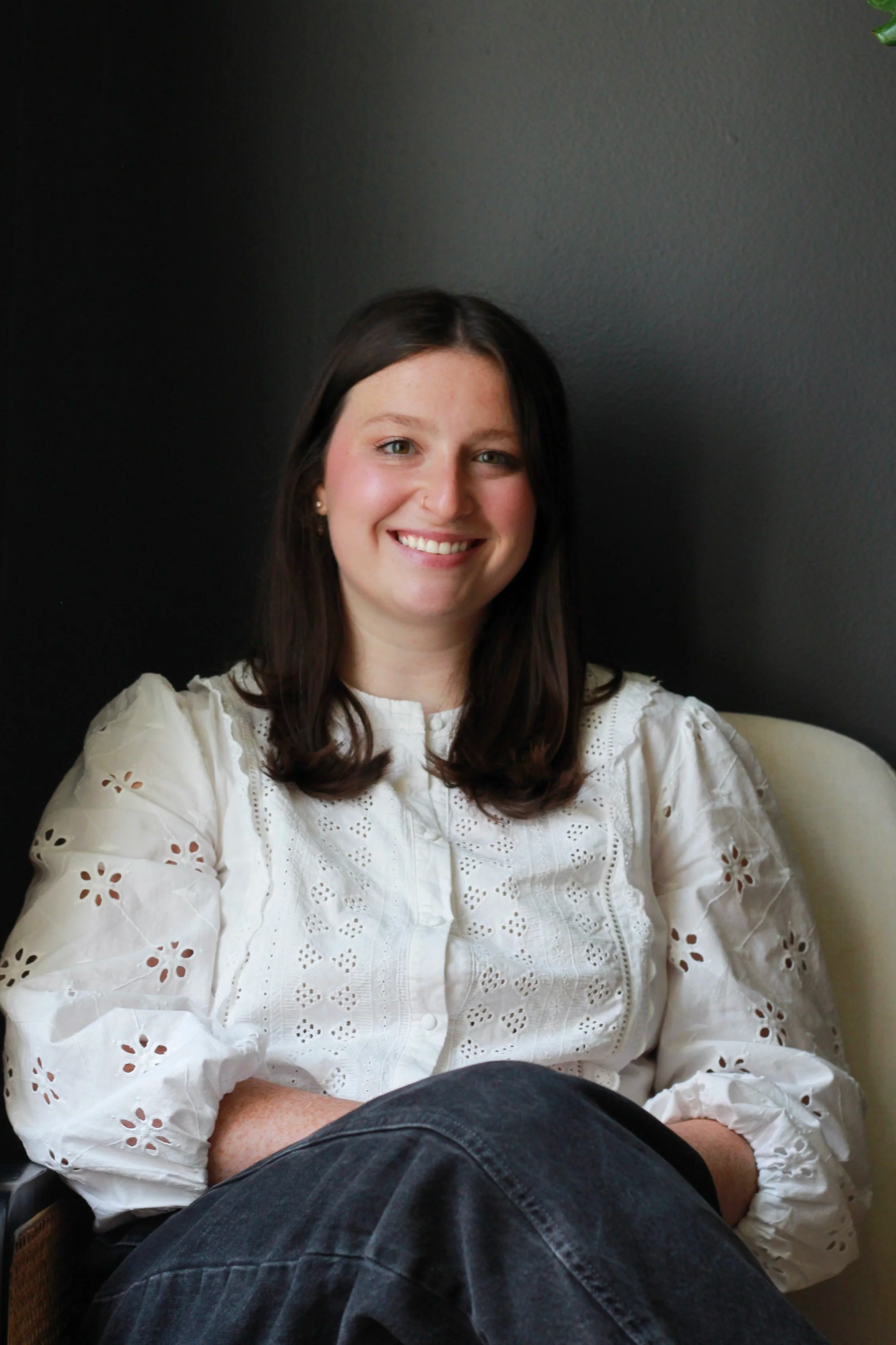 A woman with long brown hair smiling, wearing a white embroidered blouse, sitting on a beige chair against a dark wall.