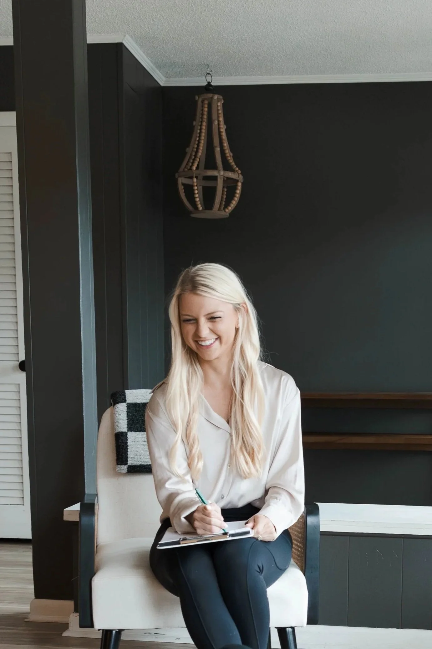 A woman with long blonde hair sitting on a white chair, smiling and holding a clipboard with a pen, in a room with dark walls and a hanging light fixture.