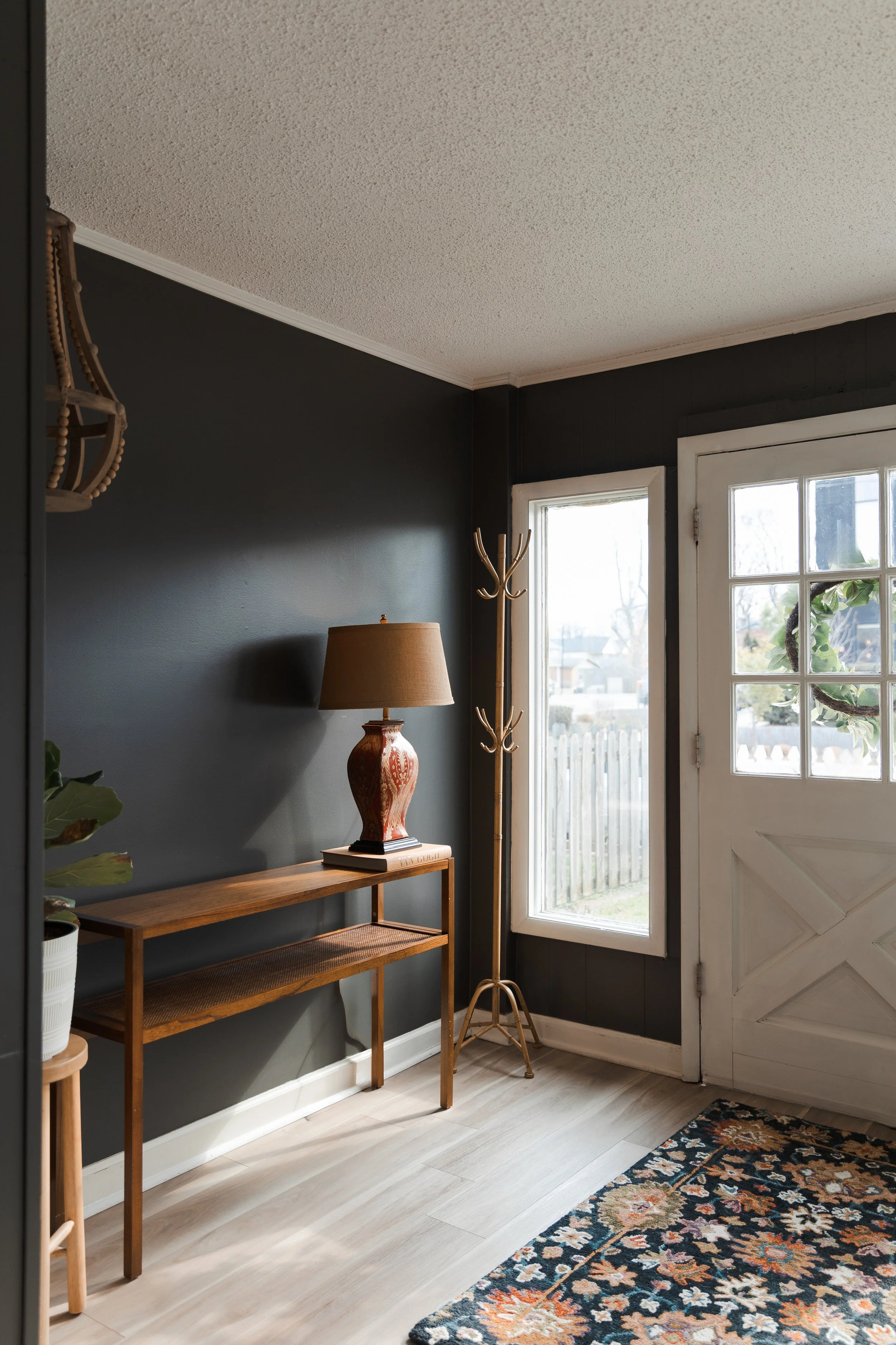 Entryway with a dark accent wall, wooden console table with a lamp, coat rack, front door with window panes, and a floral rug.