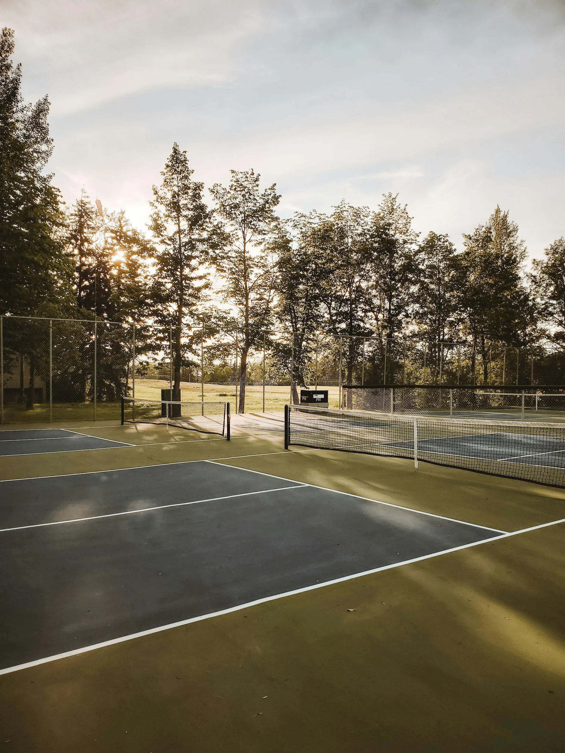 A deserted outdoor tennis court at sunset with trees in the background and a black trash can near the fence.