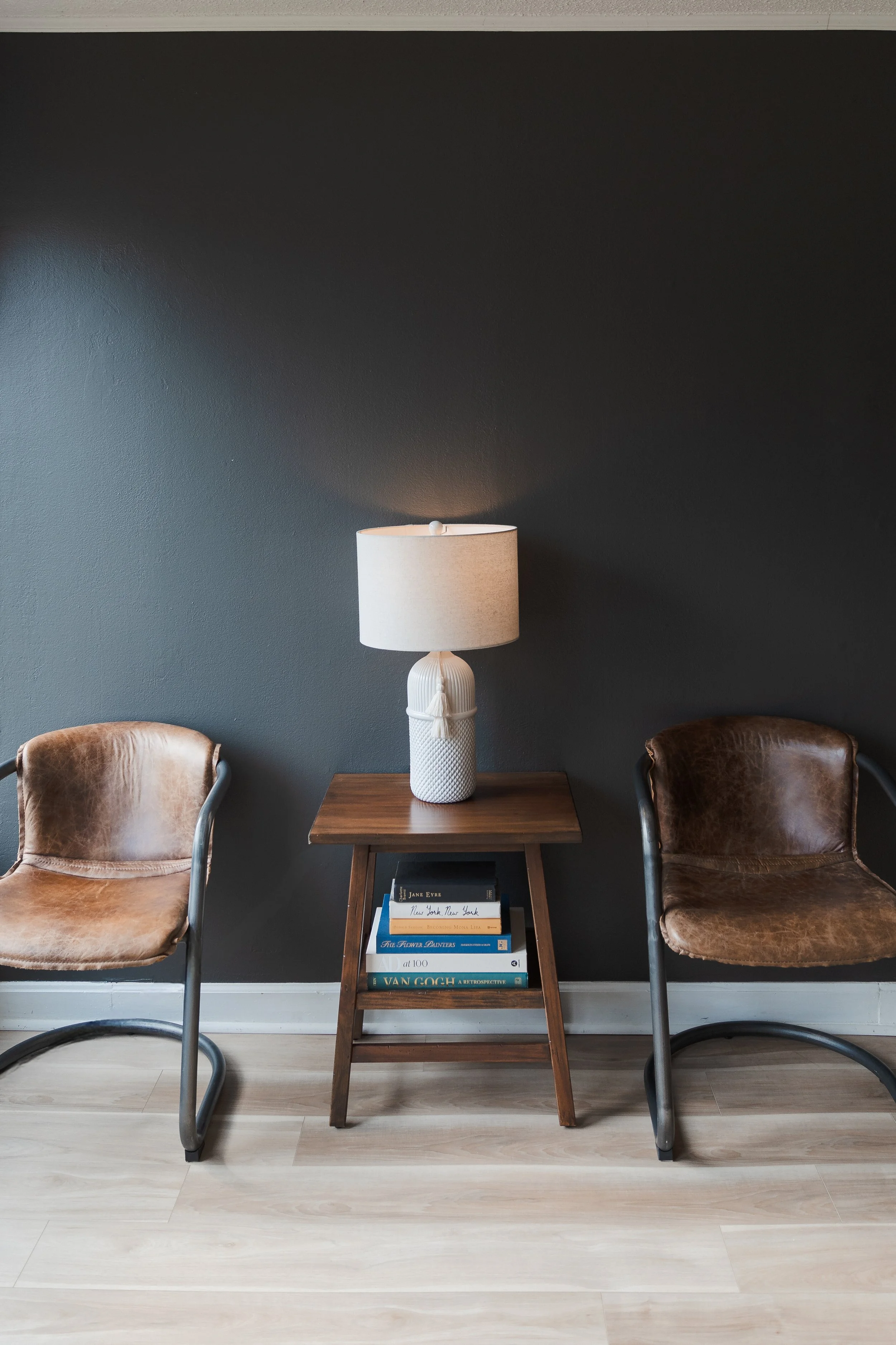 Living room with a dark gray wall, two vintage leather chairs with metal frames on either side of a small wooden side table. The table has a white ceramic lamp with a textured base, and below it are books including "New York, New York" and "VAN GOGH.