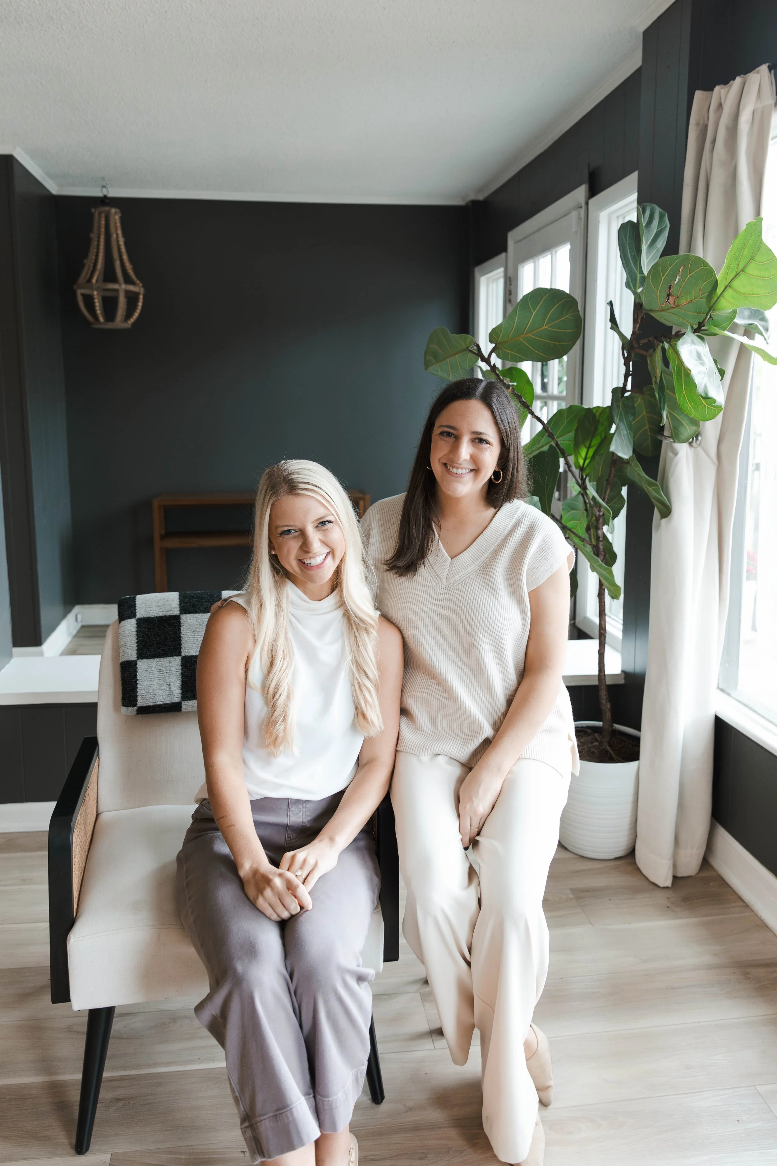 Two women sitting on a light-colored couch in a living room, smiling at the camera. One woman has long blonde hair and is wearing a sleeveless white top and gray pants. The other woman has shoulder-length dark hair and is wearing a light beige sweater and white pants. Behind them is a dark-colored wall, a large houseplant, and windows with white curtains.