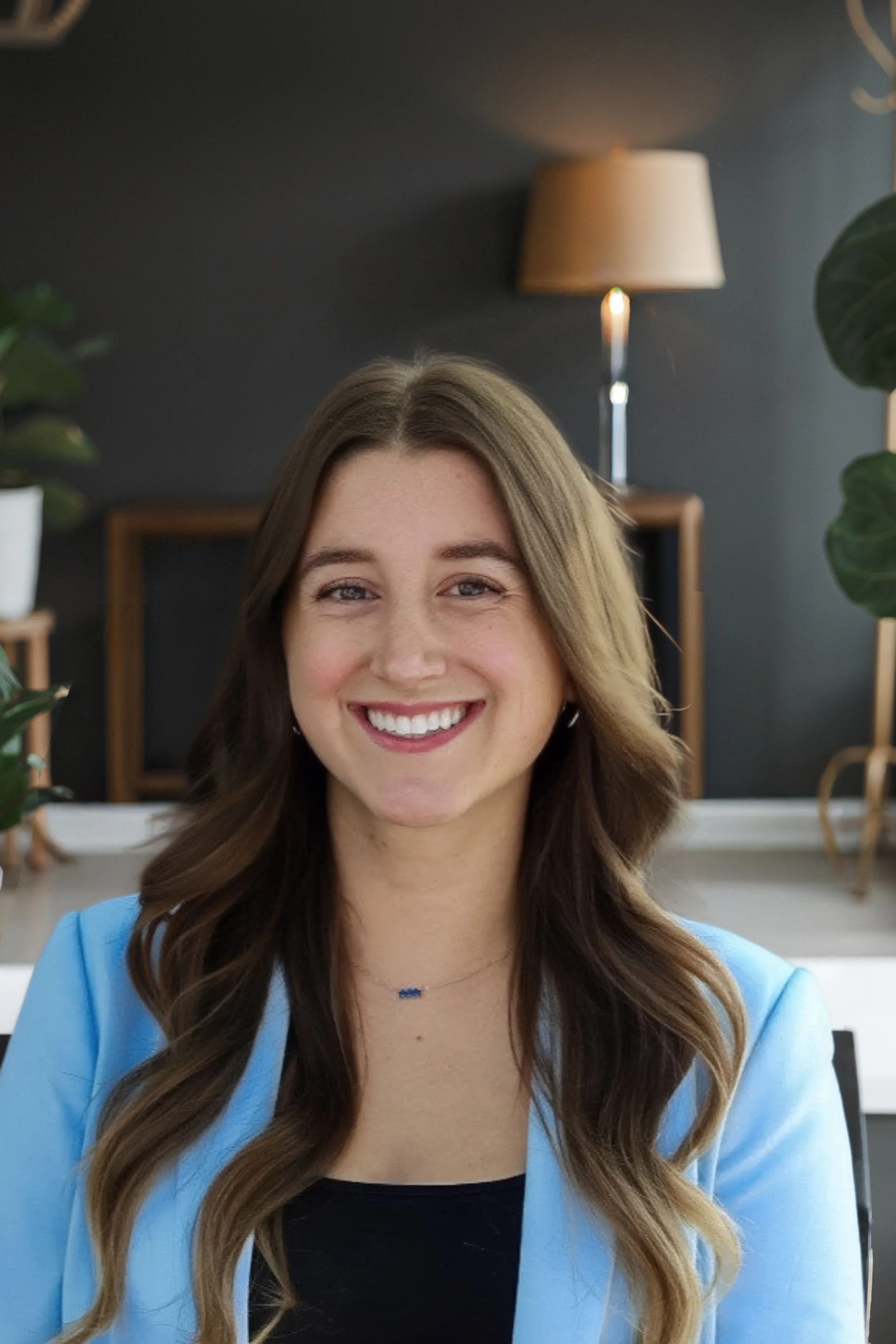 A smiling woman with long brown hair wearing a blue blazer and black top, sitting in a modern indoor setting with plants and a wall lamp in the background.