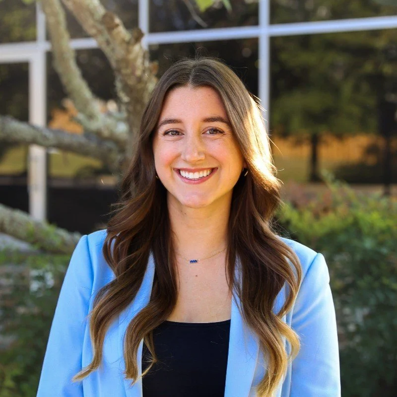 Young woman with long, wavy brown hair, smiling, wearing a black top and a light blue blazer, standing outdoors with a tree and building in the background.