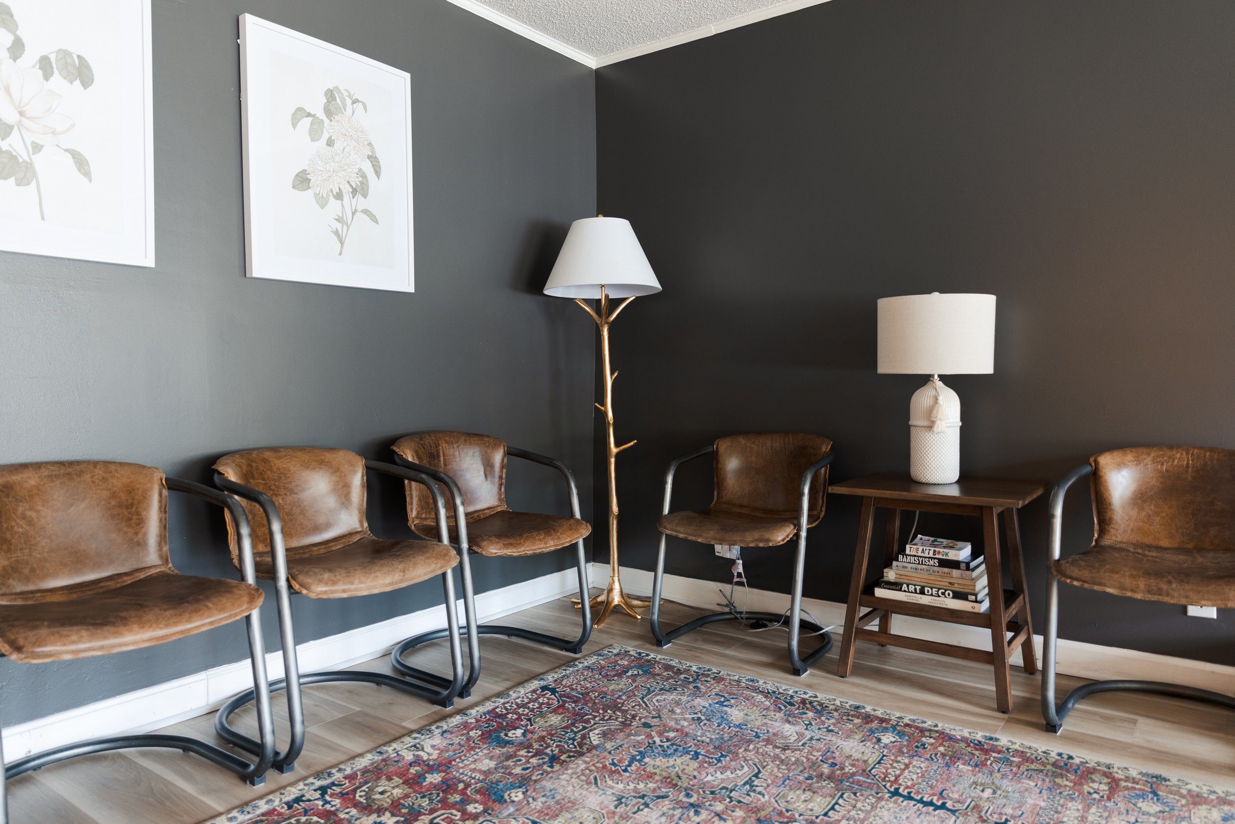 Living room with dark gray wall, four brown leather chairs with black metal frames, a wooden side table with books beneath, a white table lamp, a tall floor lamp resembling a tree branch with a white shade, and framed botanical artwork on the wall. T