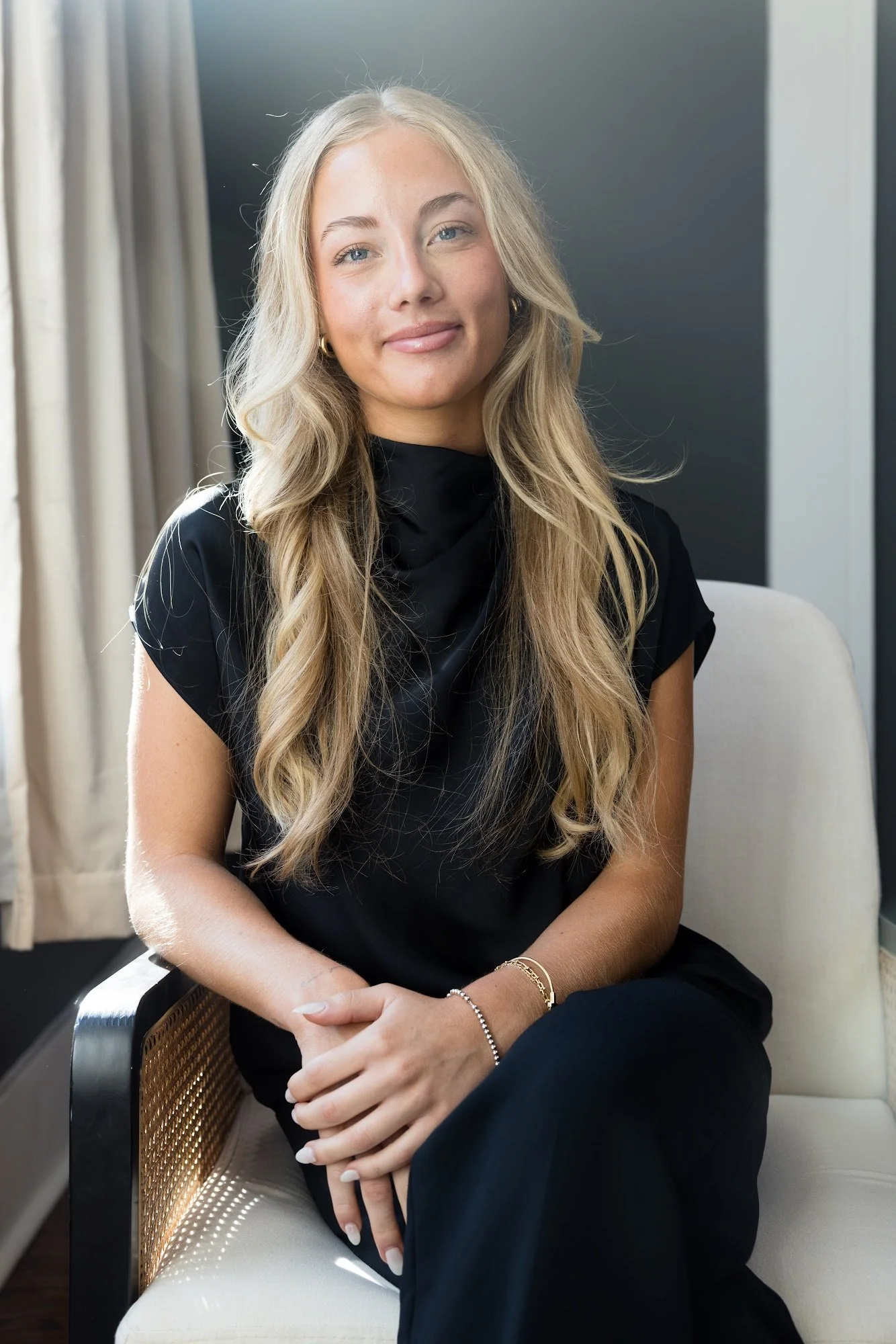 A woman with long blonde hair and light skin sitting on a chair, wearing a white top and black pants, with her hands resting on her lap in front of a dark wall.
