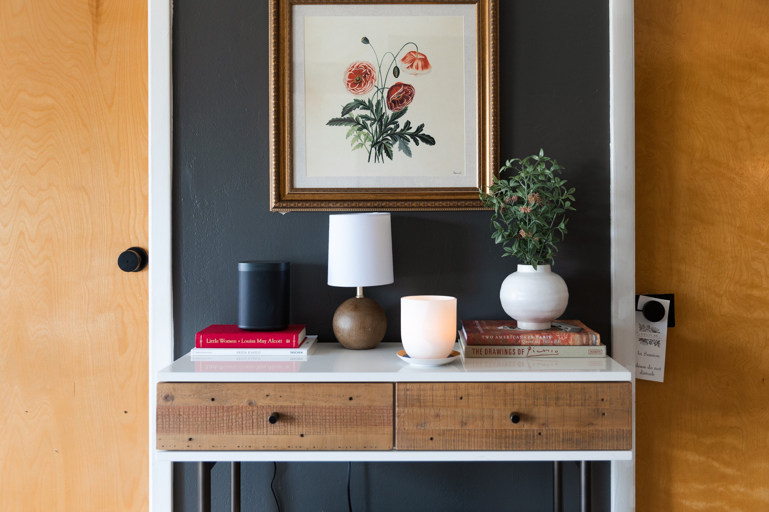 A shelf with a white surface holds a small speaker, a stack of books including one titled 'Little Women' and 'The Drawings of Piccino', a brown decorative vase, a white candle holder, and a large white vase with green leafy plants. Above the shelf, a framed botanical print of red poppies hangs on a dark gray wall.
