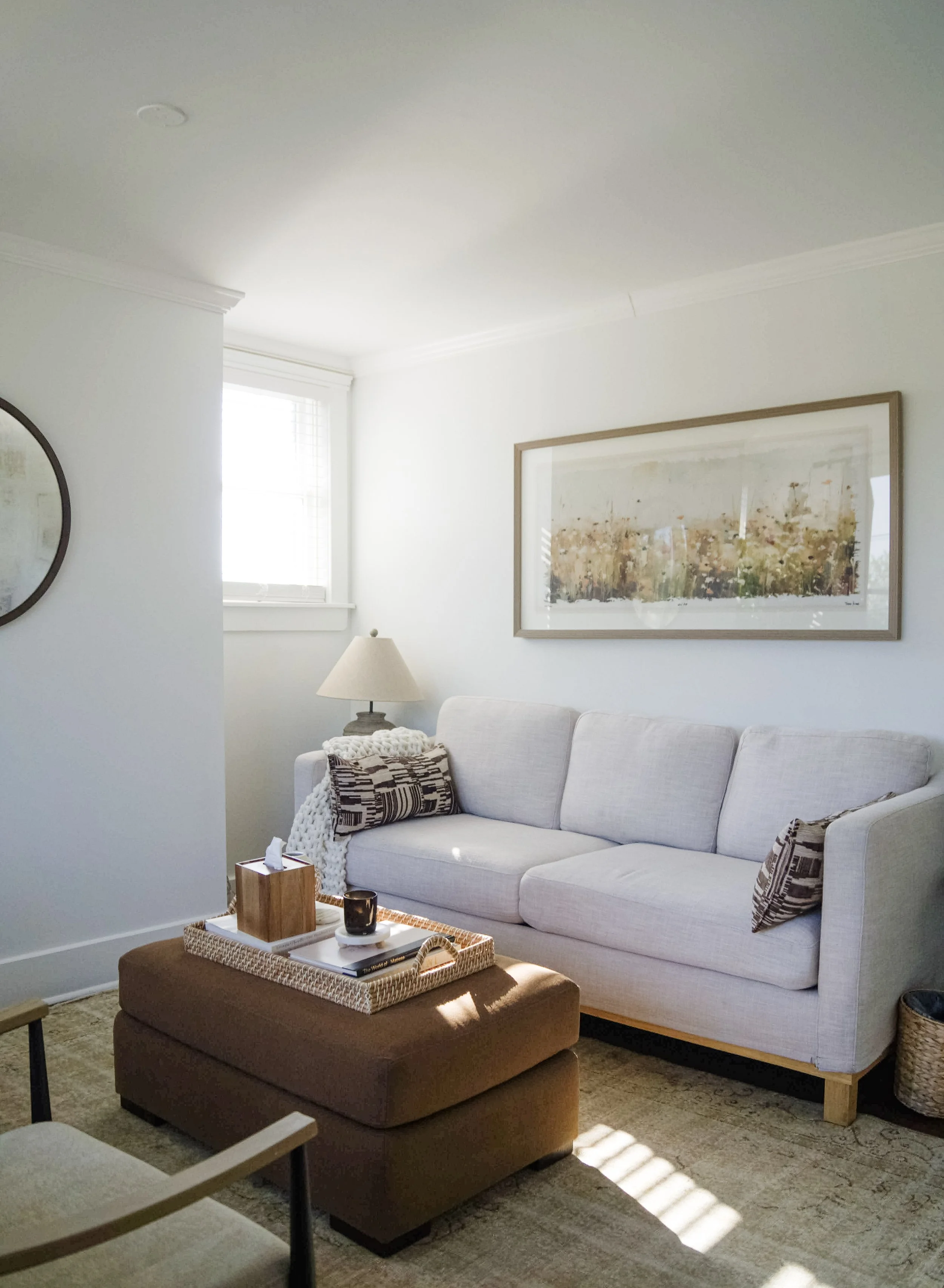 Bright living room with a white sofa, patterned pillows, a lamp, a wall art, and a brown ottoman with a tray, a mug, and a tissue box.