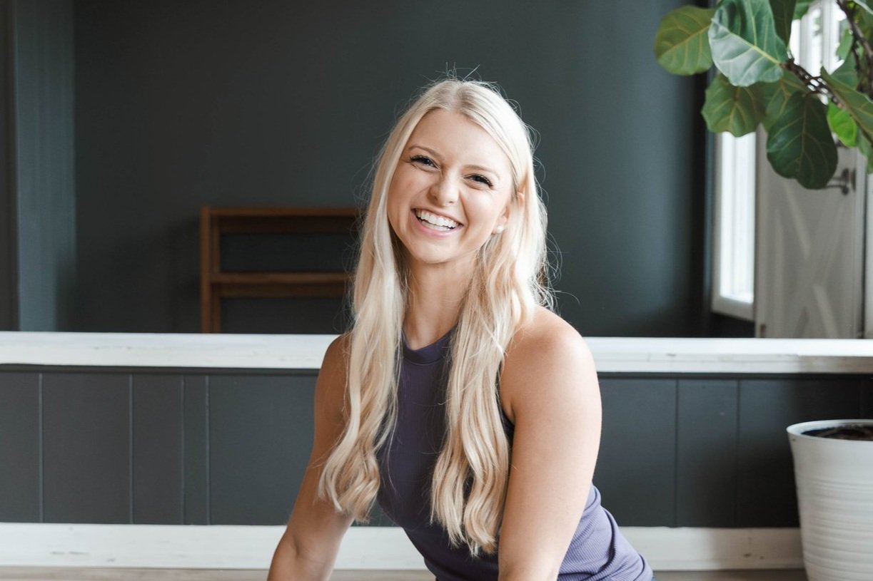 A smiling woman with long blonde hair, wearing a sleeveless gray top, sitting indoors near a potted plant and a window.