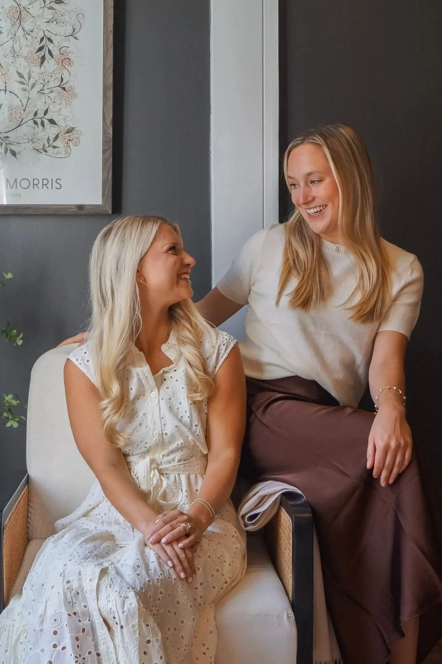 Two women with blonde hair smiling and talking in a cozy, modern room, one wearing a cream-colored top and brown skirt, the other in a white eyelet dress, sitting on a light-colored chair.