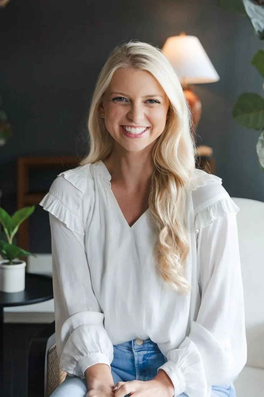 A smiling blonde woman in a white blouse sitting on a chair in a room with a dark background, a lamp, and potted plants.