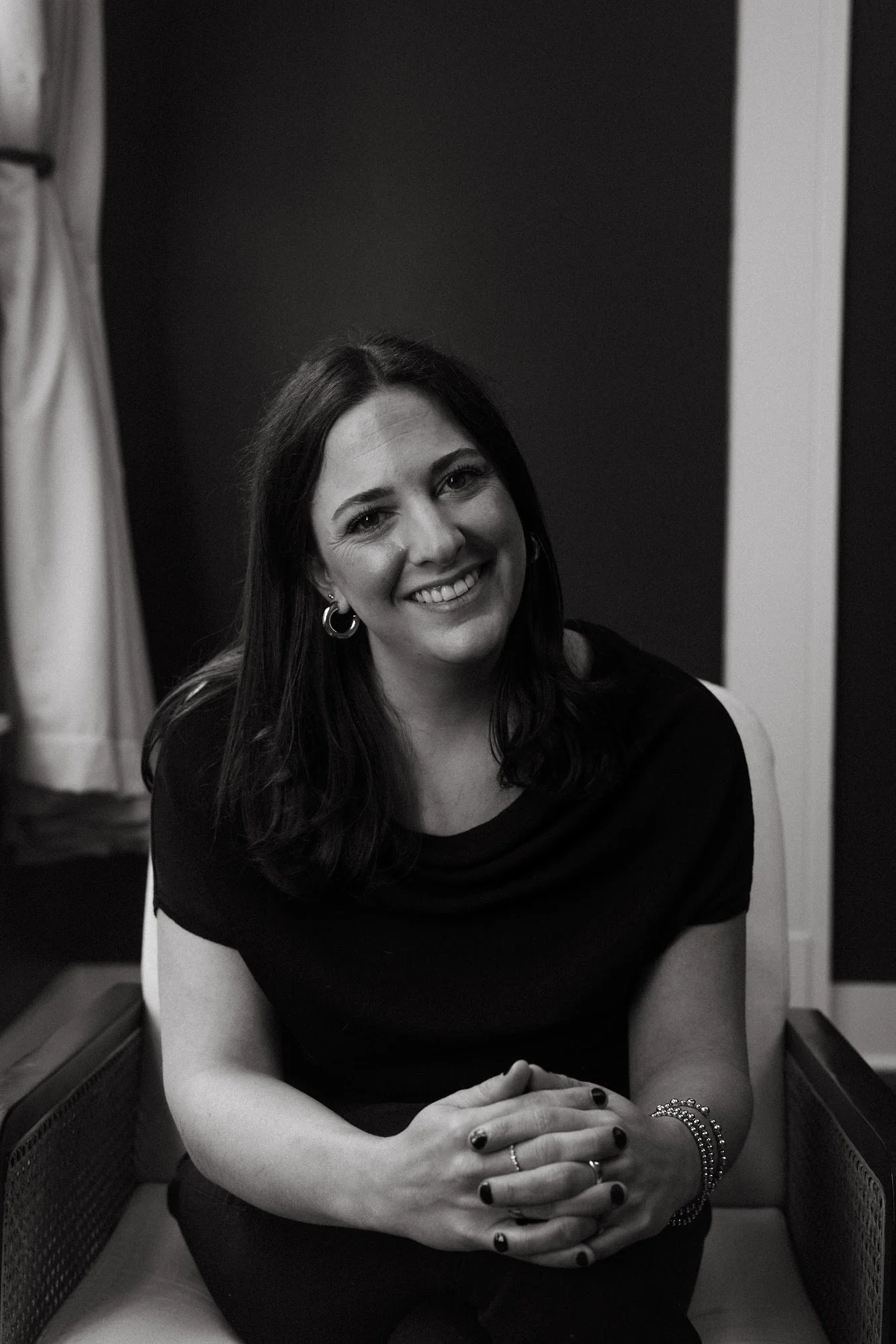 A woman with long dark hair and hoop earrings smiling while sitting in a bright room with potted plants, a round beige lampshade, and dark gray walls.