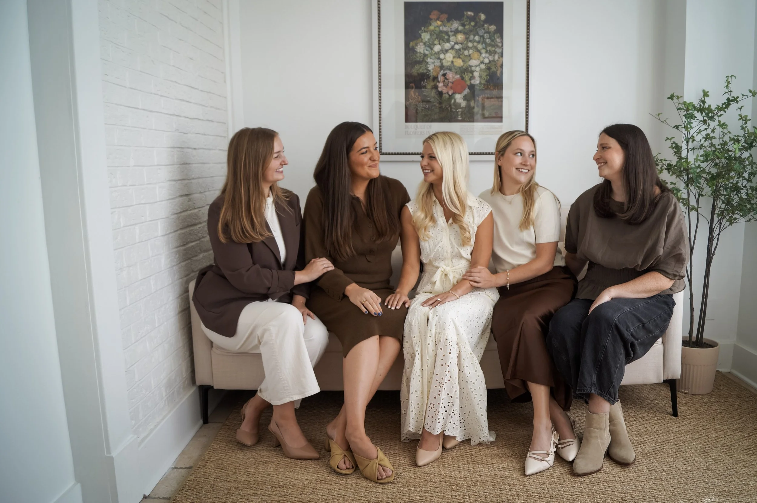 Five women sitting on a beige bench in a cozy, well-lit room, smiling and engaging with each other. They are dressed in stylish, semi-formal attire, with some wearing heels and others in boots. The room has a white brick wall on the left, a framed floral artwork above them, and a tall green potted plant on the right.