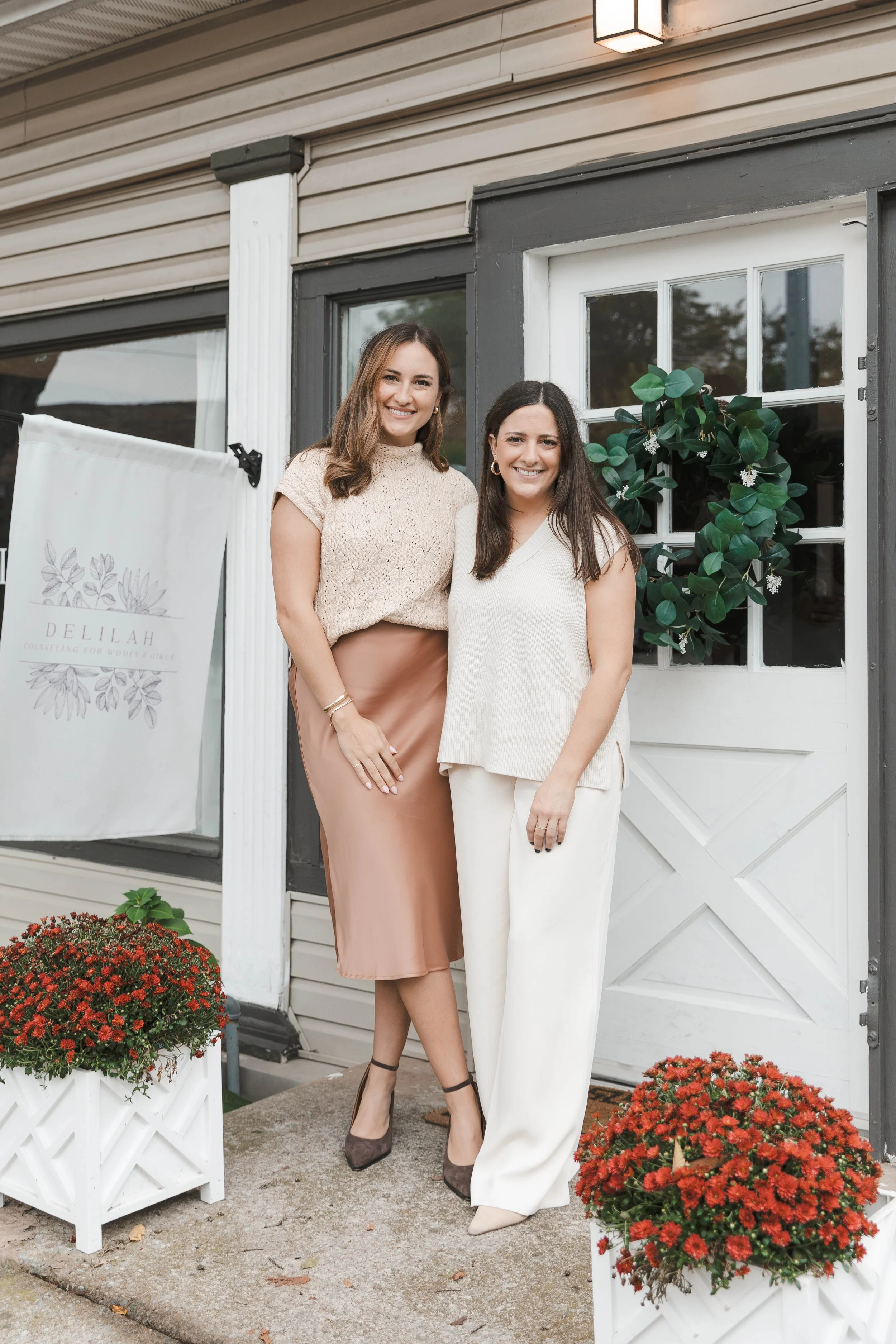 Two women standing outside in front of a building, smiling at the camera. One woman is wearing a beige top and a satin skirt, the other a cream-colored sleeveless top and white pants. There are red chrysanthemums in white planters on either side of them and greenery on the door behind them.