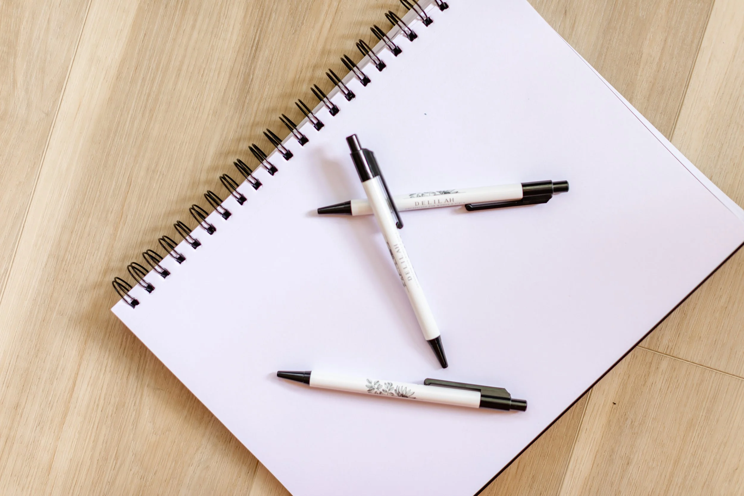 Open blank spiral notebook on a wooden surface with four black-and-white pens arranged on top.