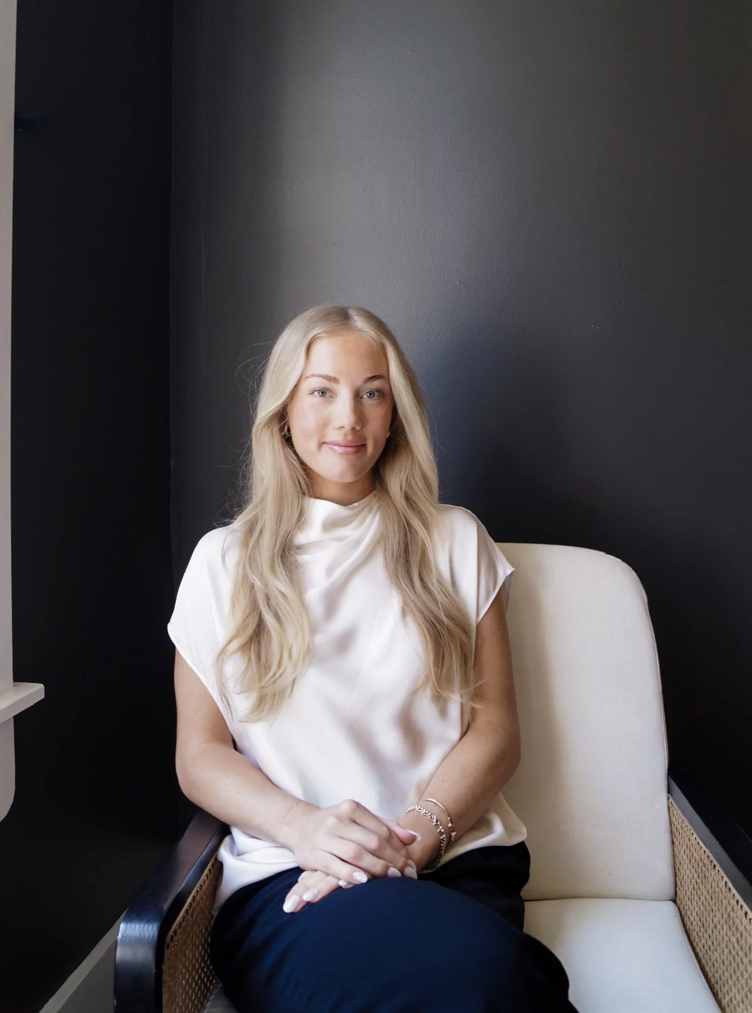 A young woman with long blonde hair, wearing a white silk top, is sitting on a beige chair with rattan sides against a dark gray wall. She has a smile, and her hands are resting on her lap, wearing bracelets.