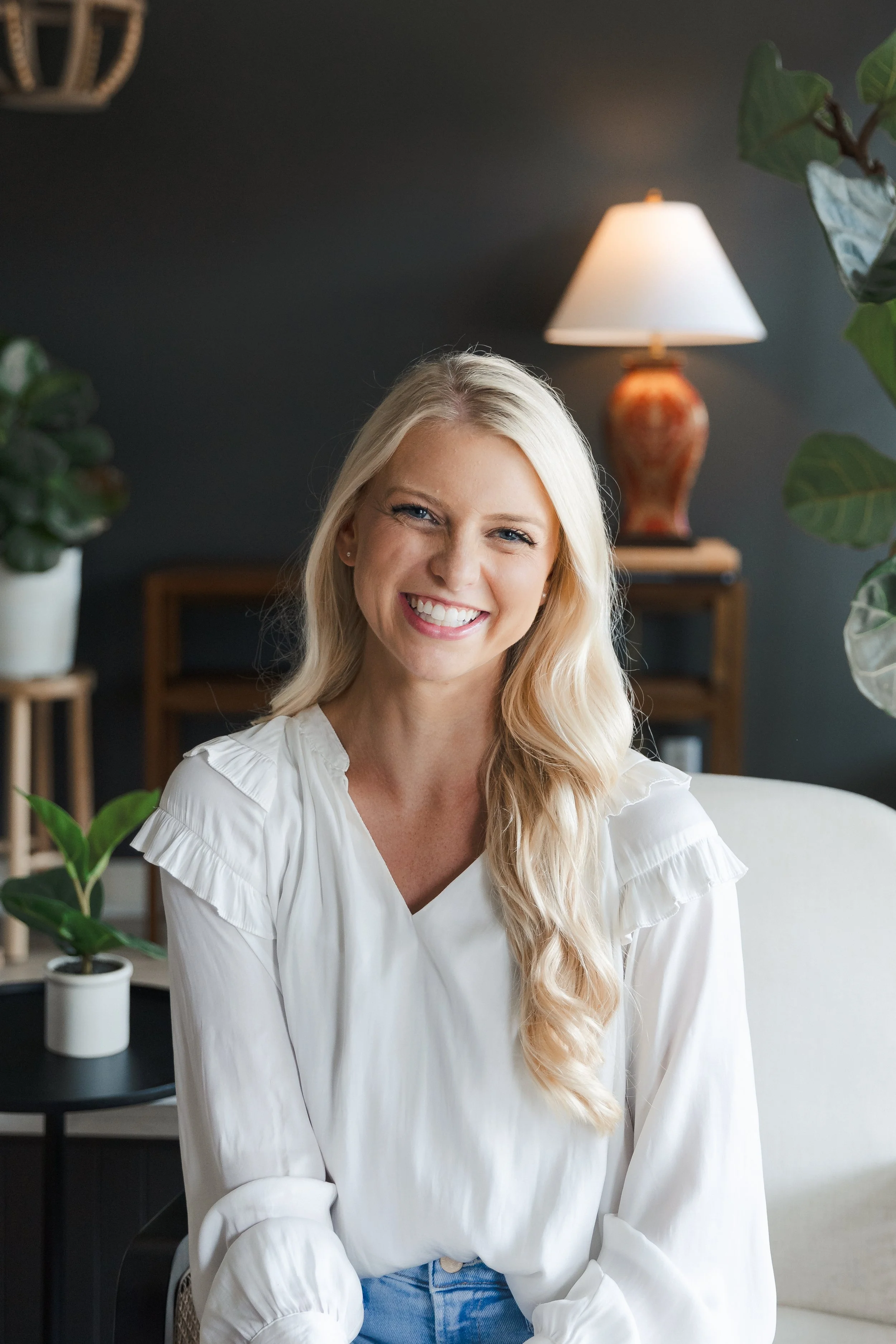 A smiling blonde woman in a white blouse sitting indoors with plants and a lamp in the background.